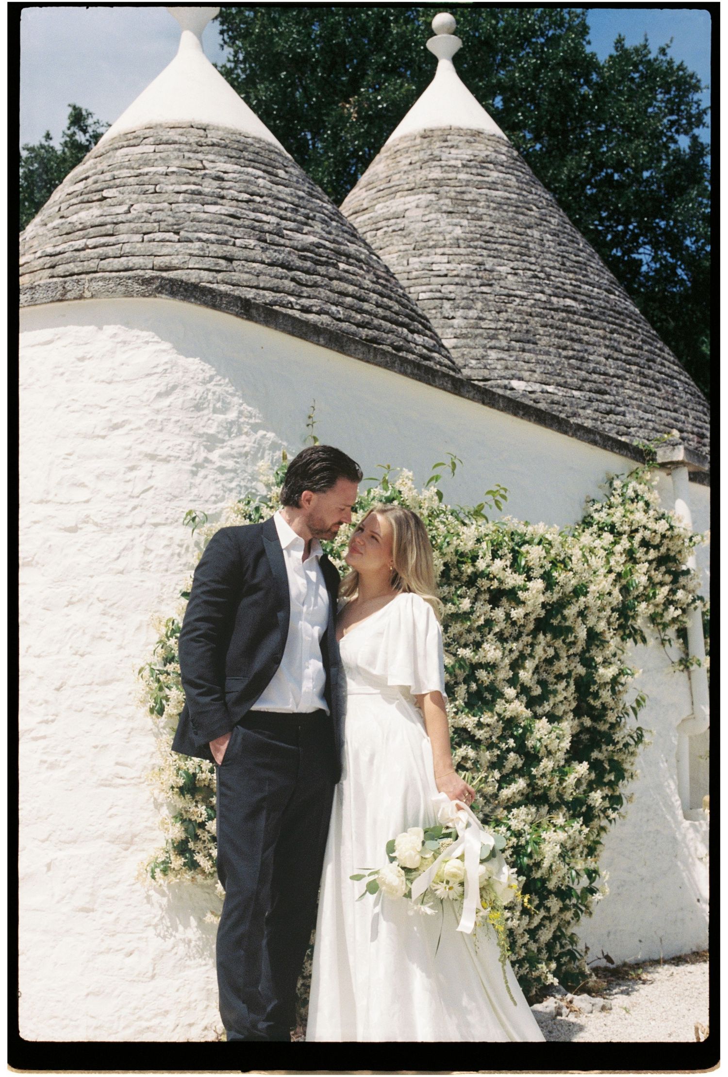 Wedding couple embracing before white trulli buildings with climbing white flowers in Puglia.