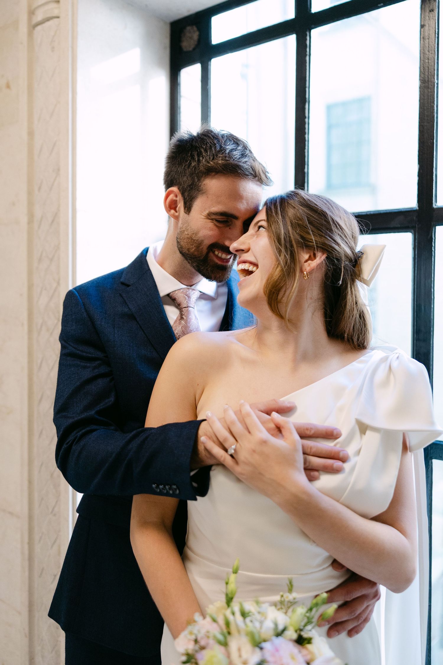 A romantic moment between a couple in formal attire embracing and laughing by large windows at The Old Marylebone Town Hall.