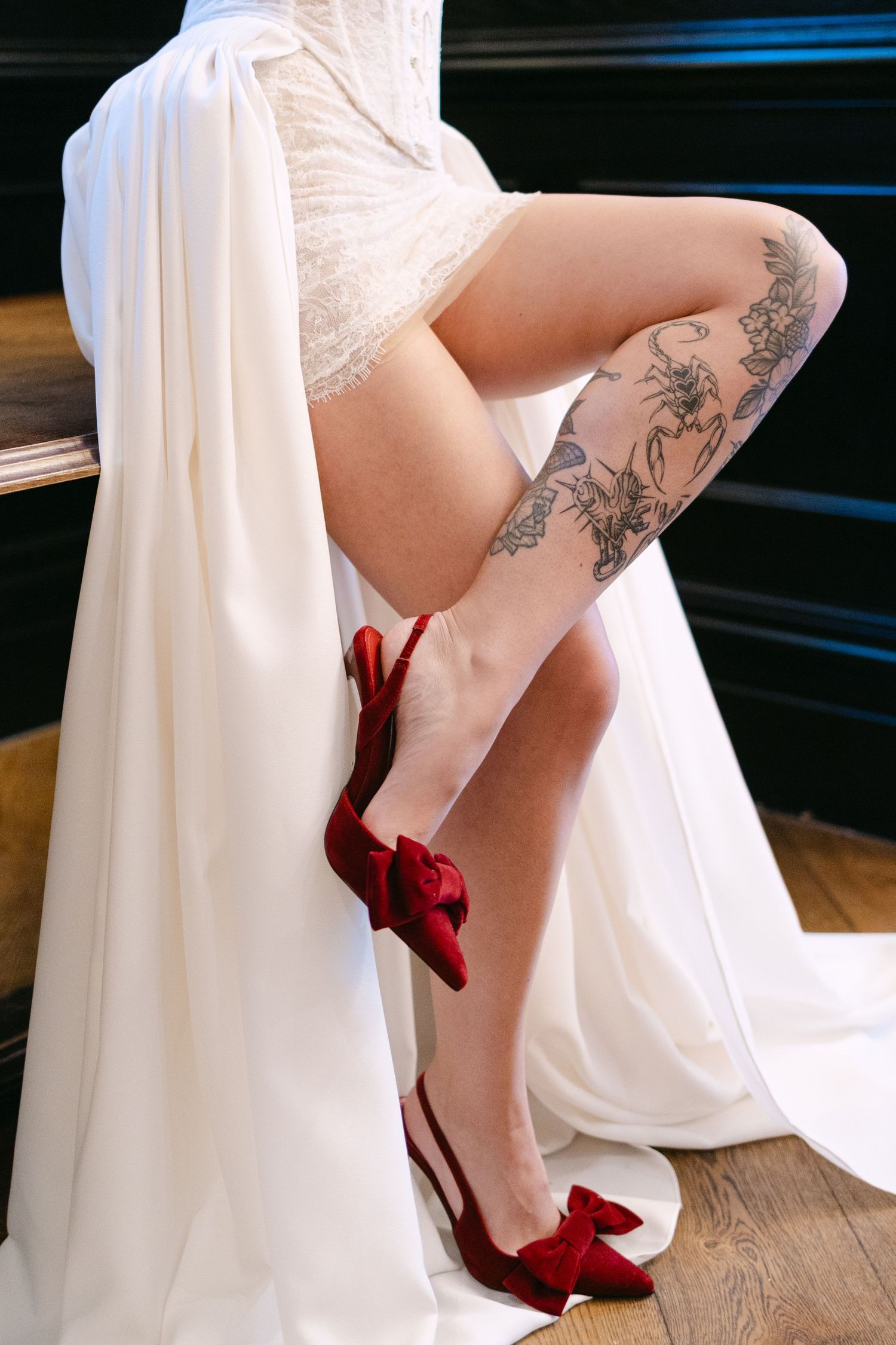 Bride in white wedding dress with red velvet bow heels and floral tattoo on leg, seated elegantly.