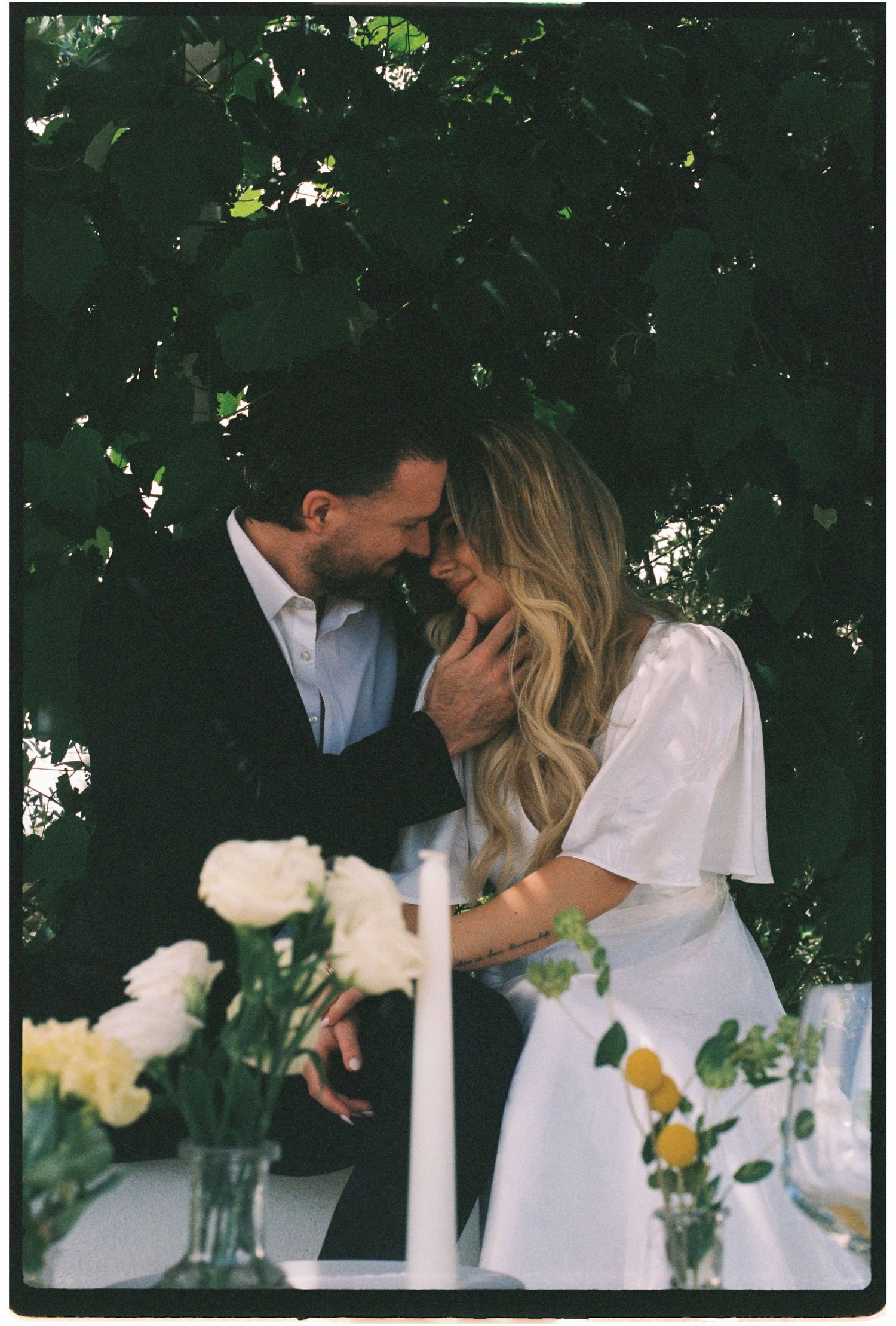 Couple shares an intimate moment leaning close together at a floral table beneath green foliage.