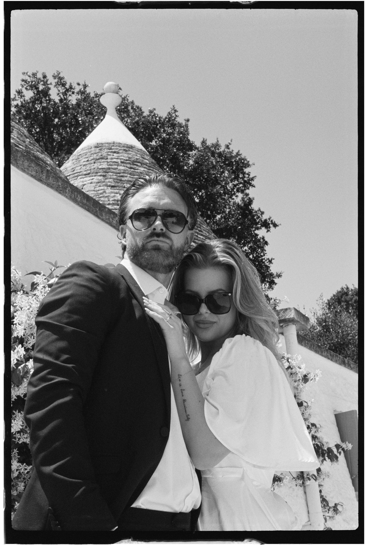 Black and white photo of a stylish couple posing outdoors near a trullo building in Italy.