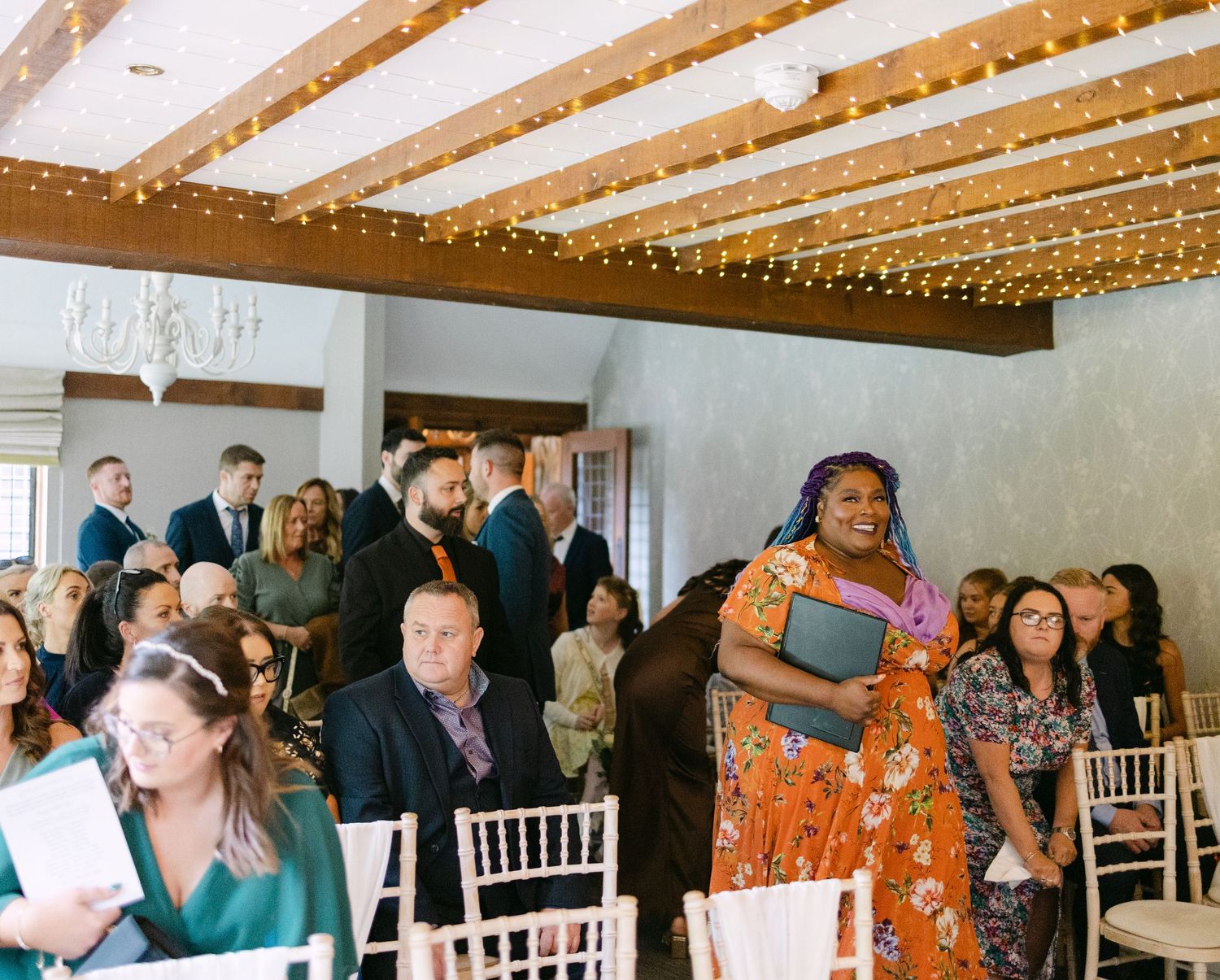Wedding guests mingle and socialize during a reception under wooden beams with string lights.