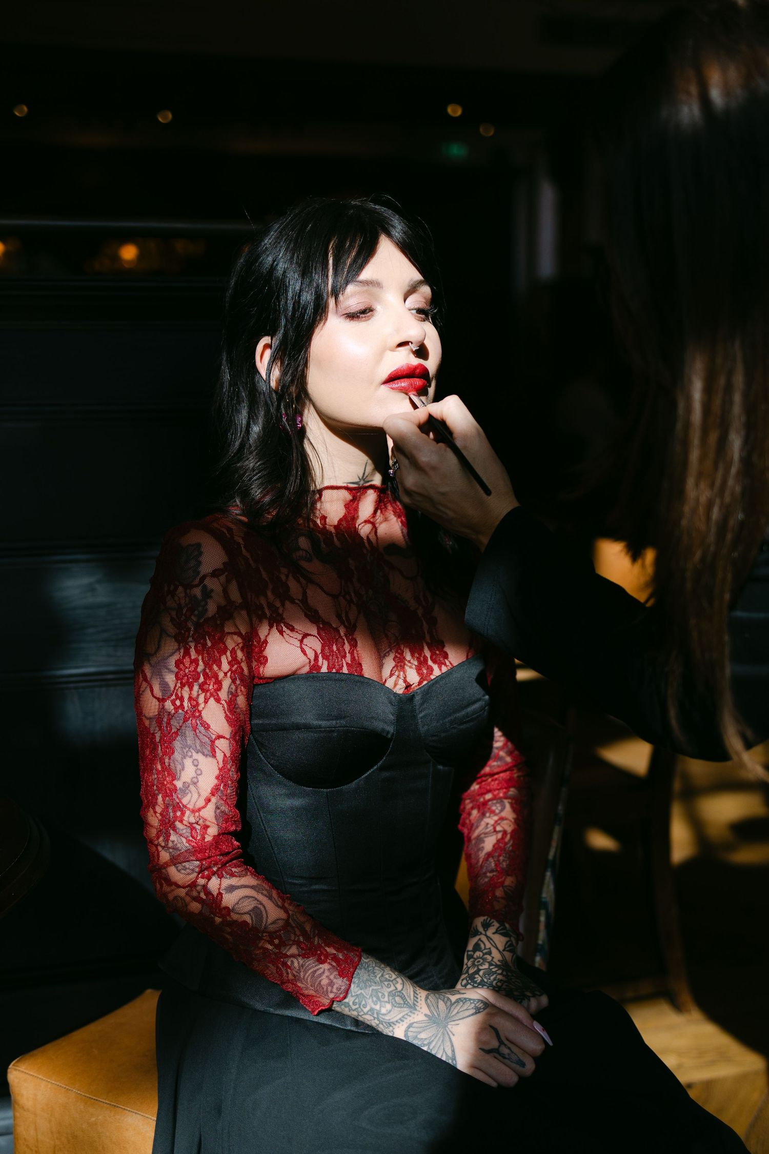 A woman in a black corset dress with red lace sleeves gets lipstick applied in a dimly lit setting.