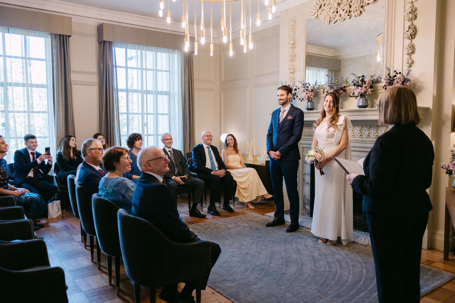 Wedding guests gather in an elegant room with chandeliers and large windows for an indoor ceremony.
