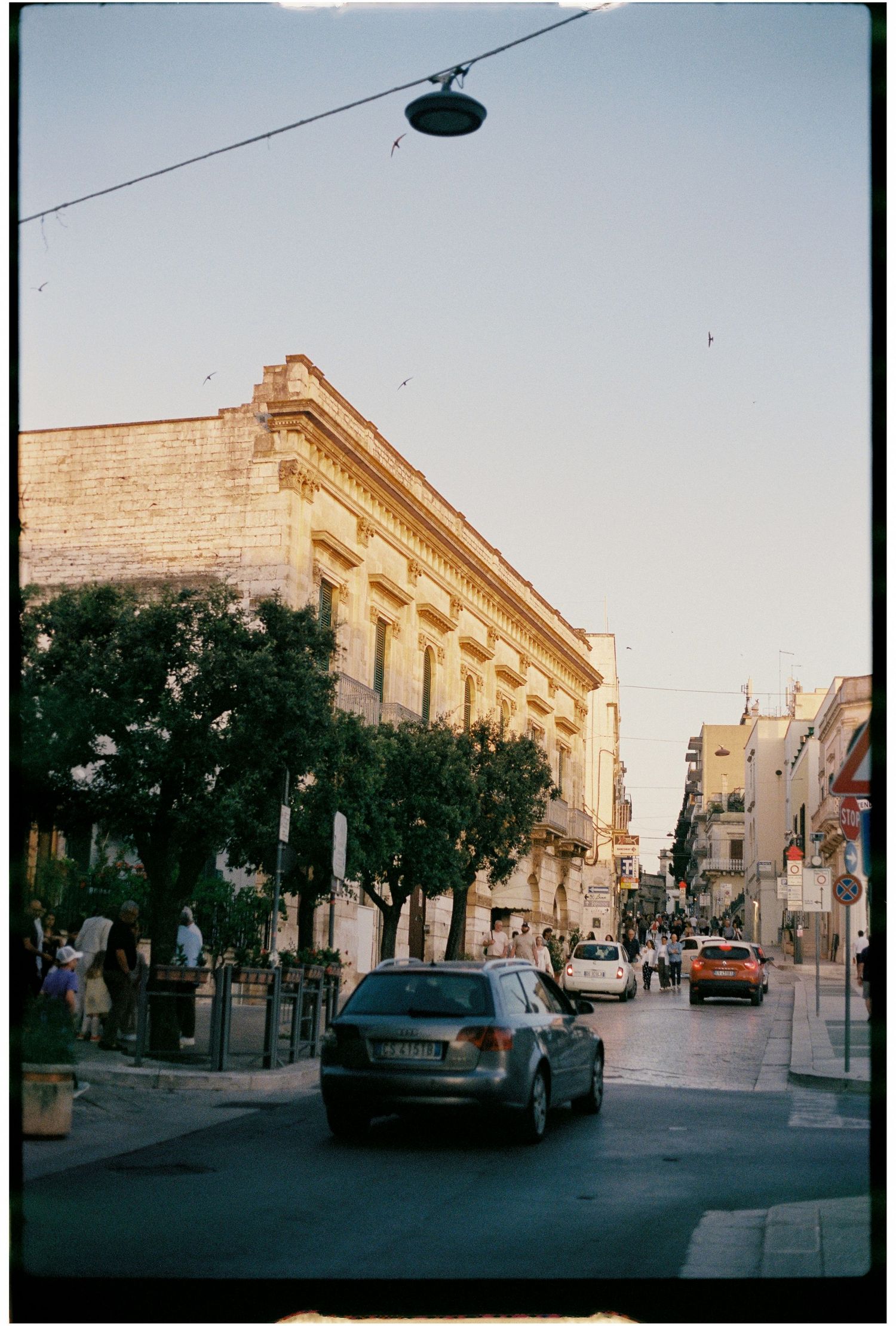 Sunlit street in a historic Italian town with stone buildings, parked cars, and pedestrians at dusk.