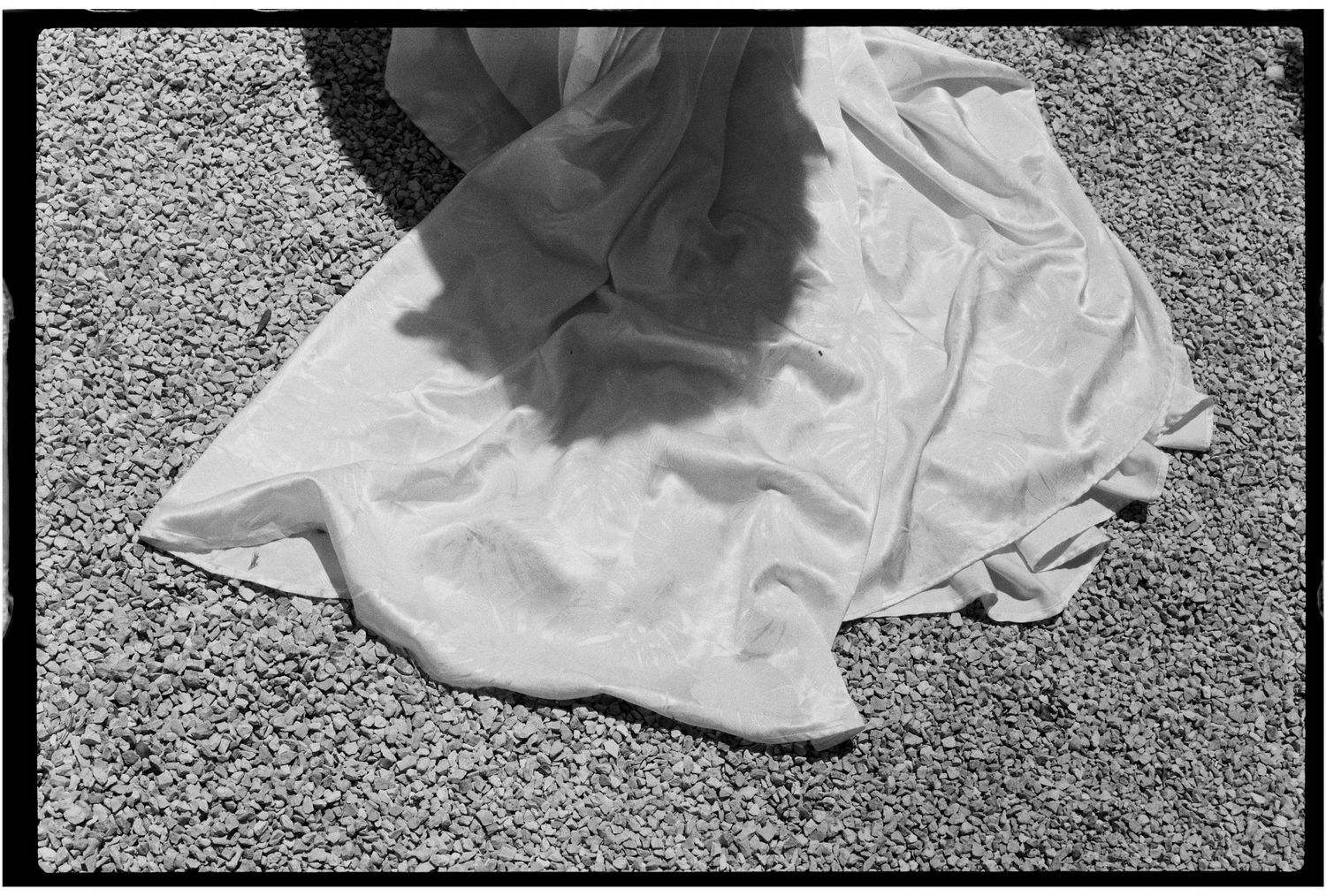 Black and white photo of a flowing white wedding dress spread on gravel ground.