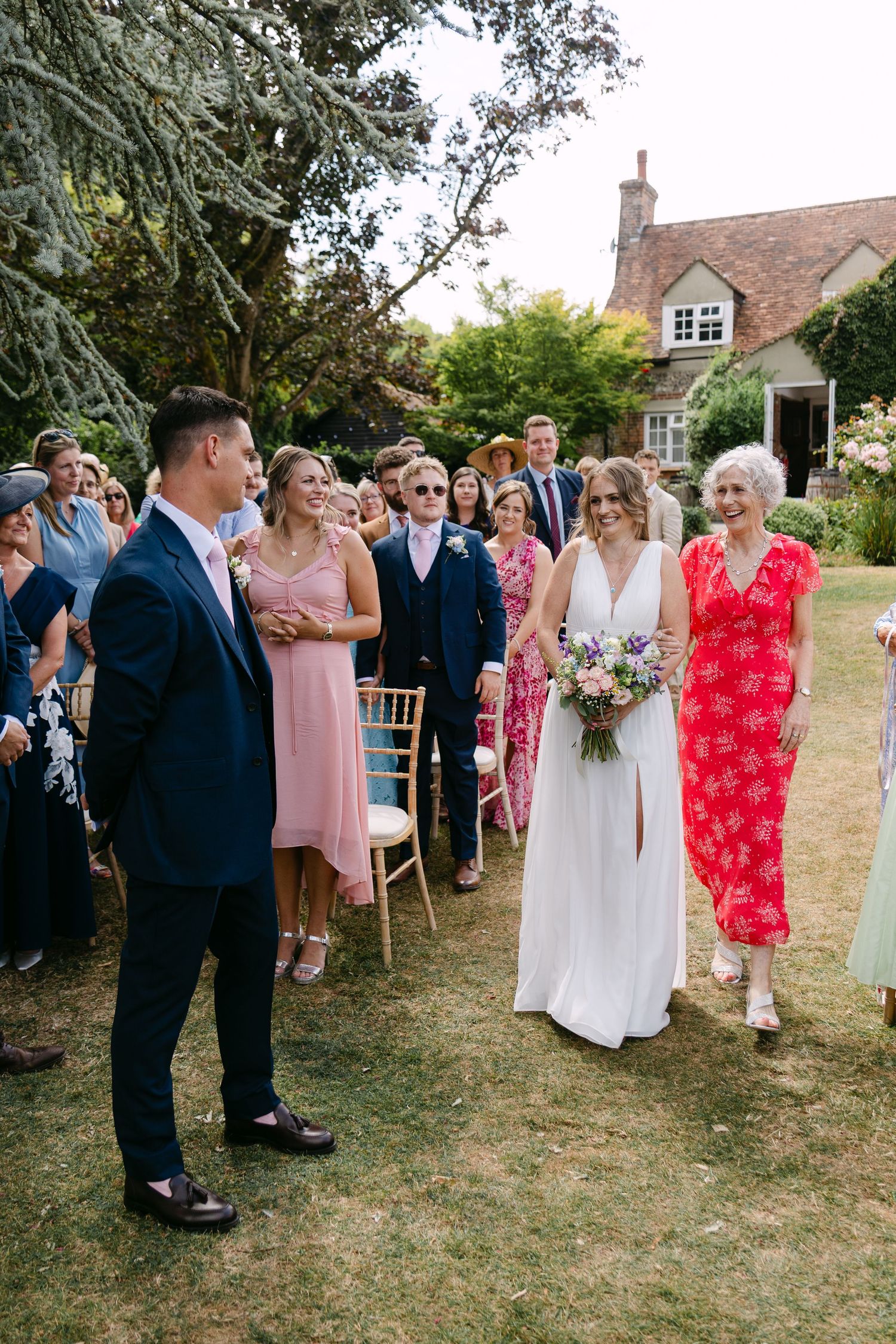 Bride walks down the aisle in a white gown holding a bouquet at an outdoor garden wedding ceremony.