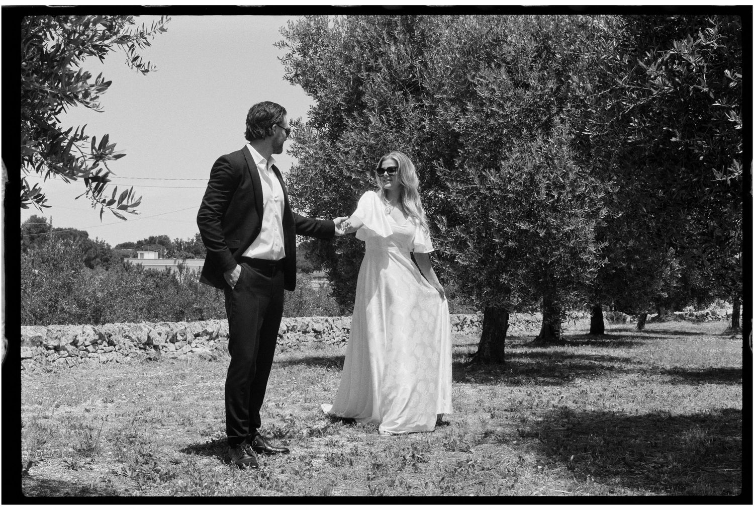 Couple in wedding attire holding hands among olive trees in a black and white outdoor photo.