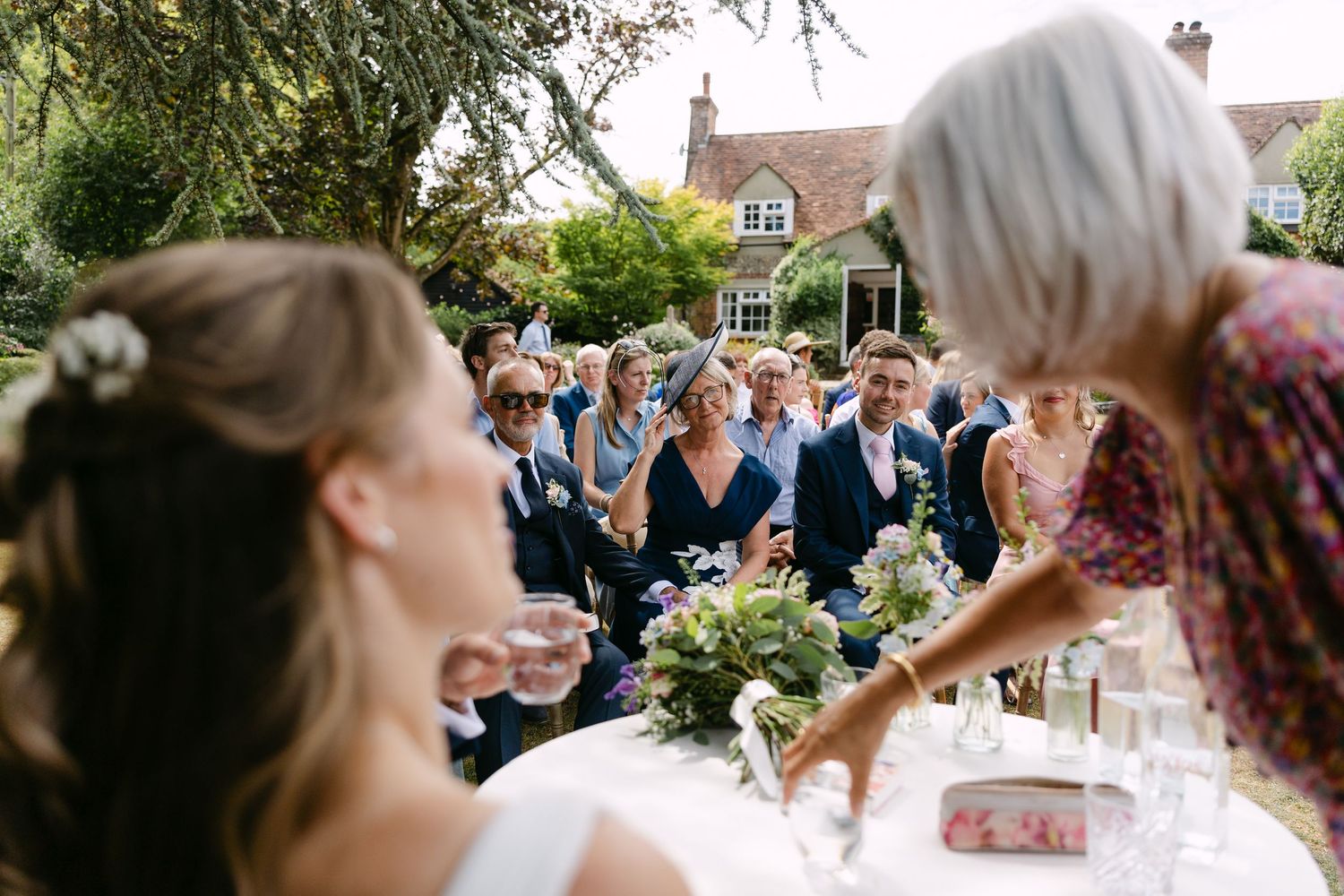 Outdoor wedding reception with guests in blue attire gathered in a garden, bride in foreground with floral arrangements.