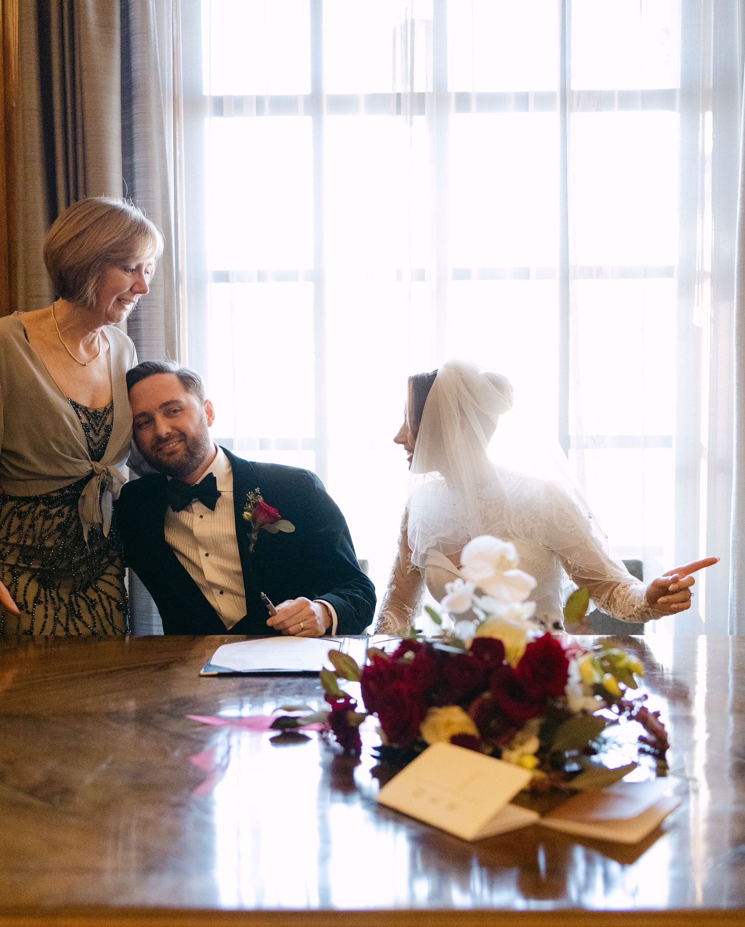 Wedding party gathered around table with red floral centerpiece during document signing.