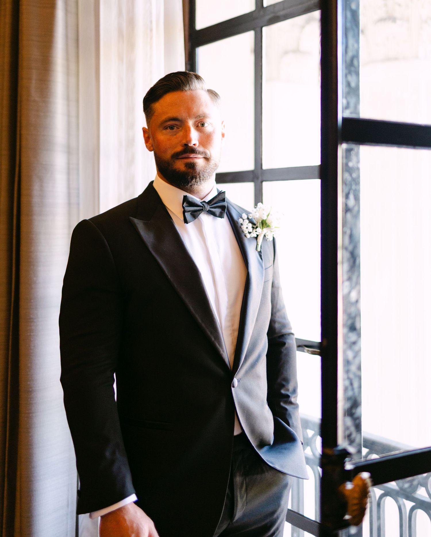 A formal portrait of a groom in a black tuxedo and bow tie by a window.