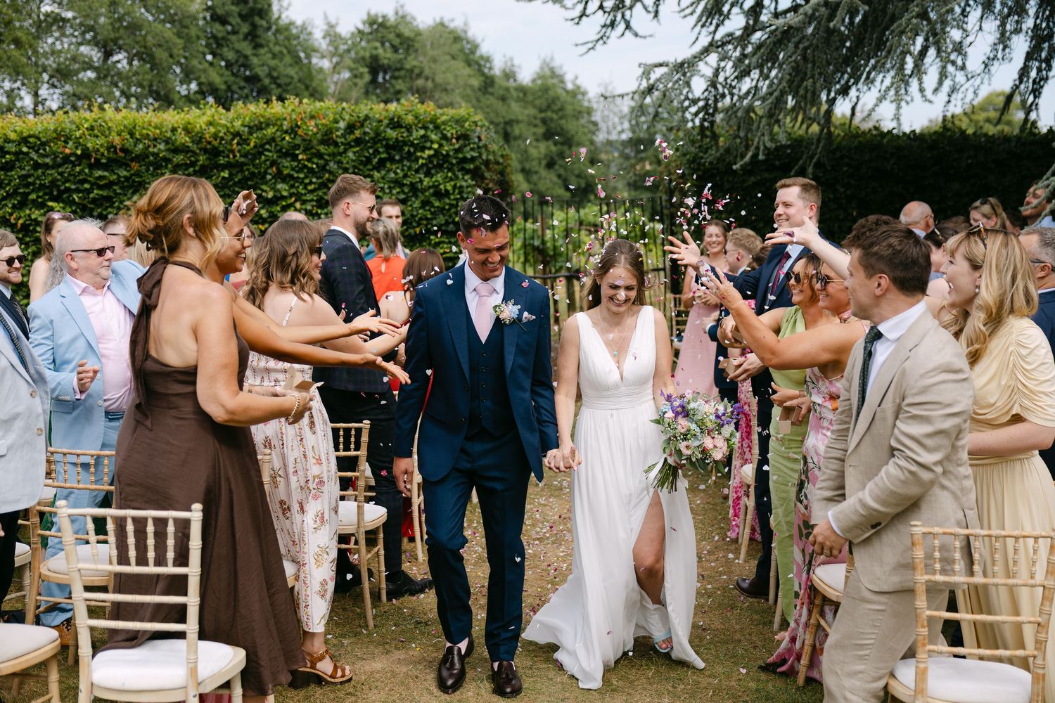 Newlyweds walk down the aisle as guests throw confetti at an outdoor garden wedding ceremony.