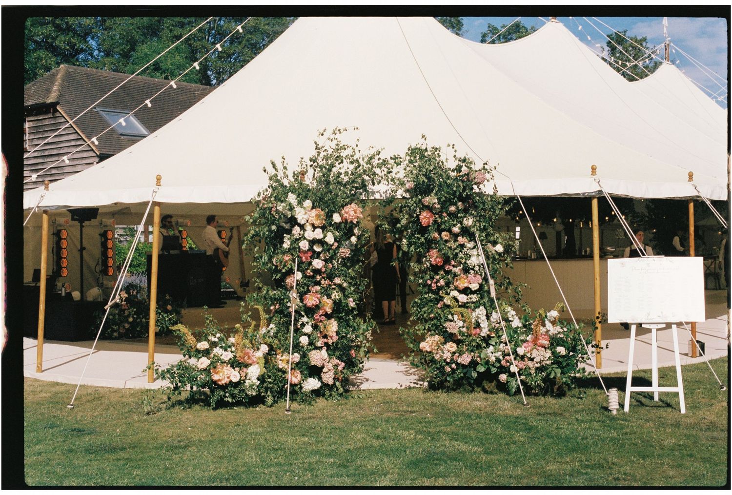 Elegant outdoor wedding tent entrance adorned with a lush floral arch of pink and peach blooms on green lawn.