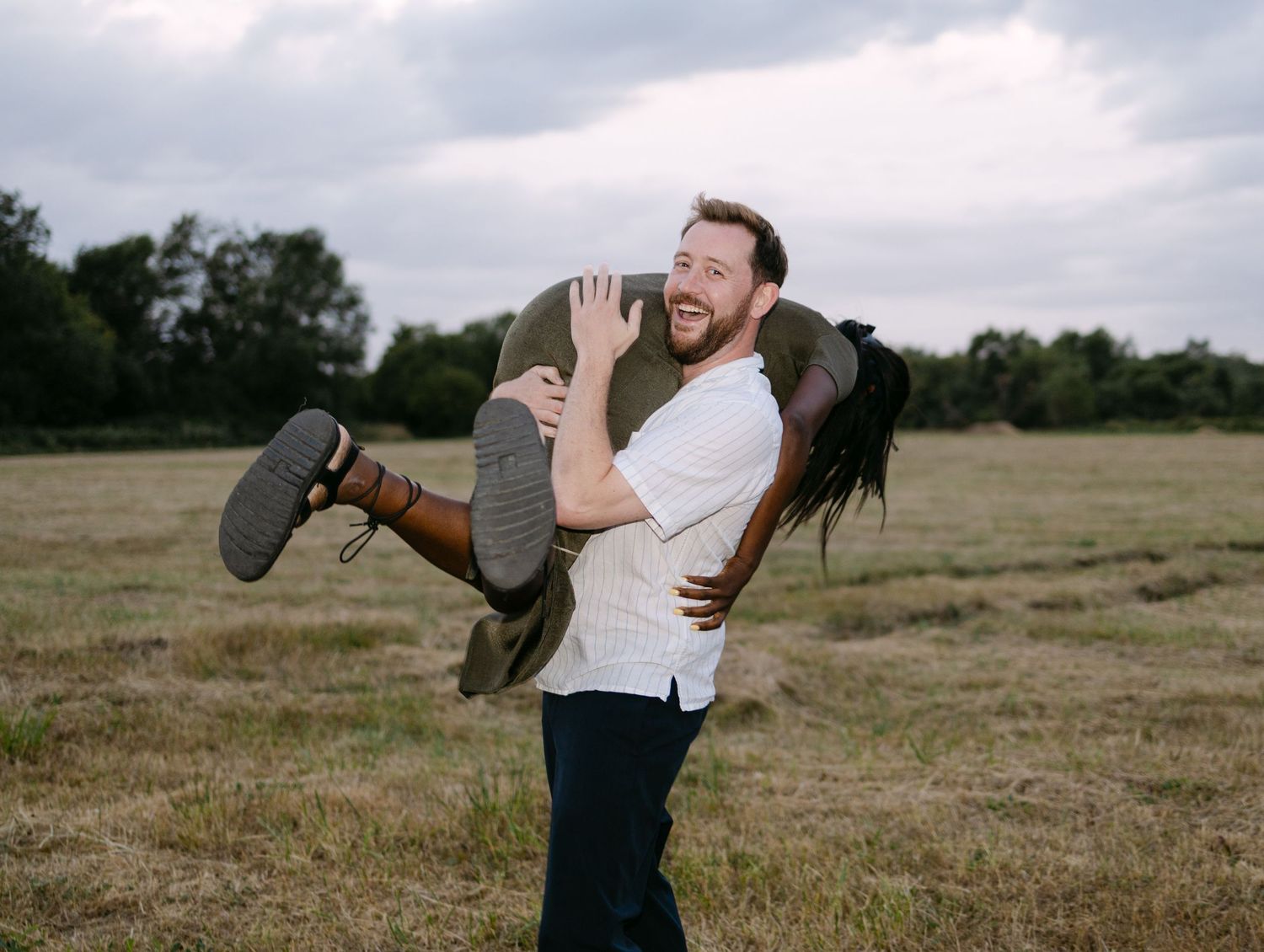 A couple embraces and spins playfully in a grassy meadow during a romantic outdoor photoshoot.