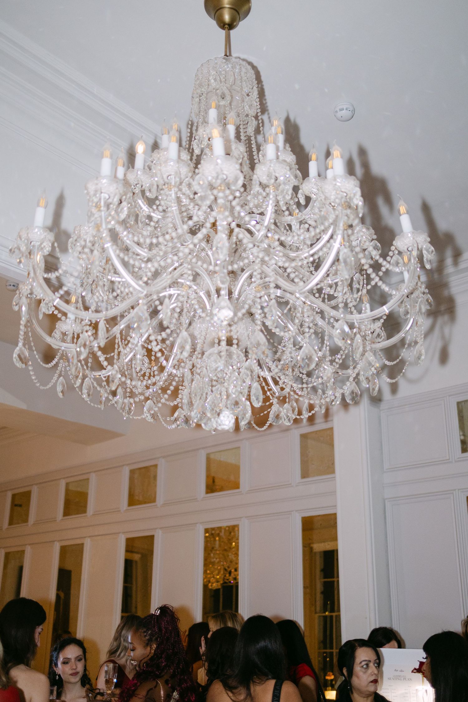 Elegant crystal chandelier with candle lights hanging in a white ballroom above a crowd of guests.