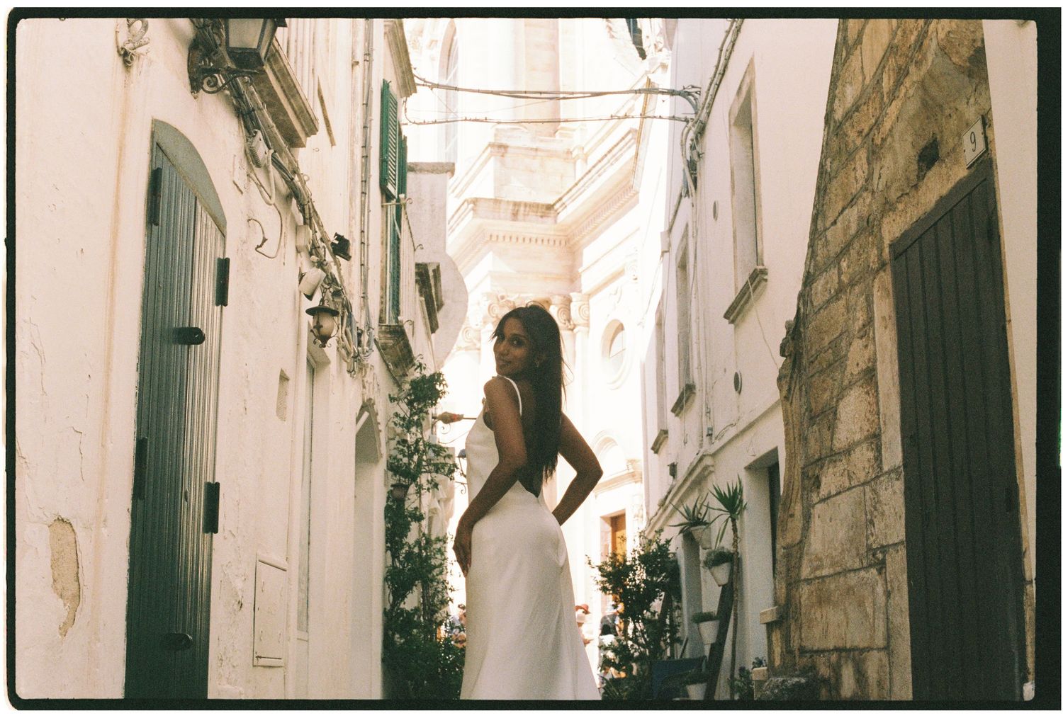 Woman in elegant white backless dress posing in a narrow sunlit Italian alleyway.