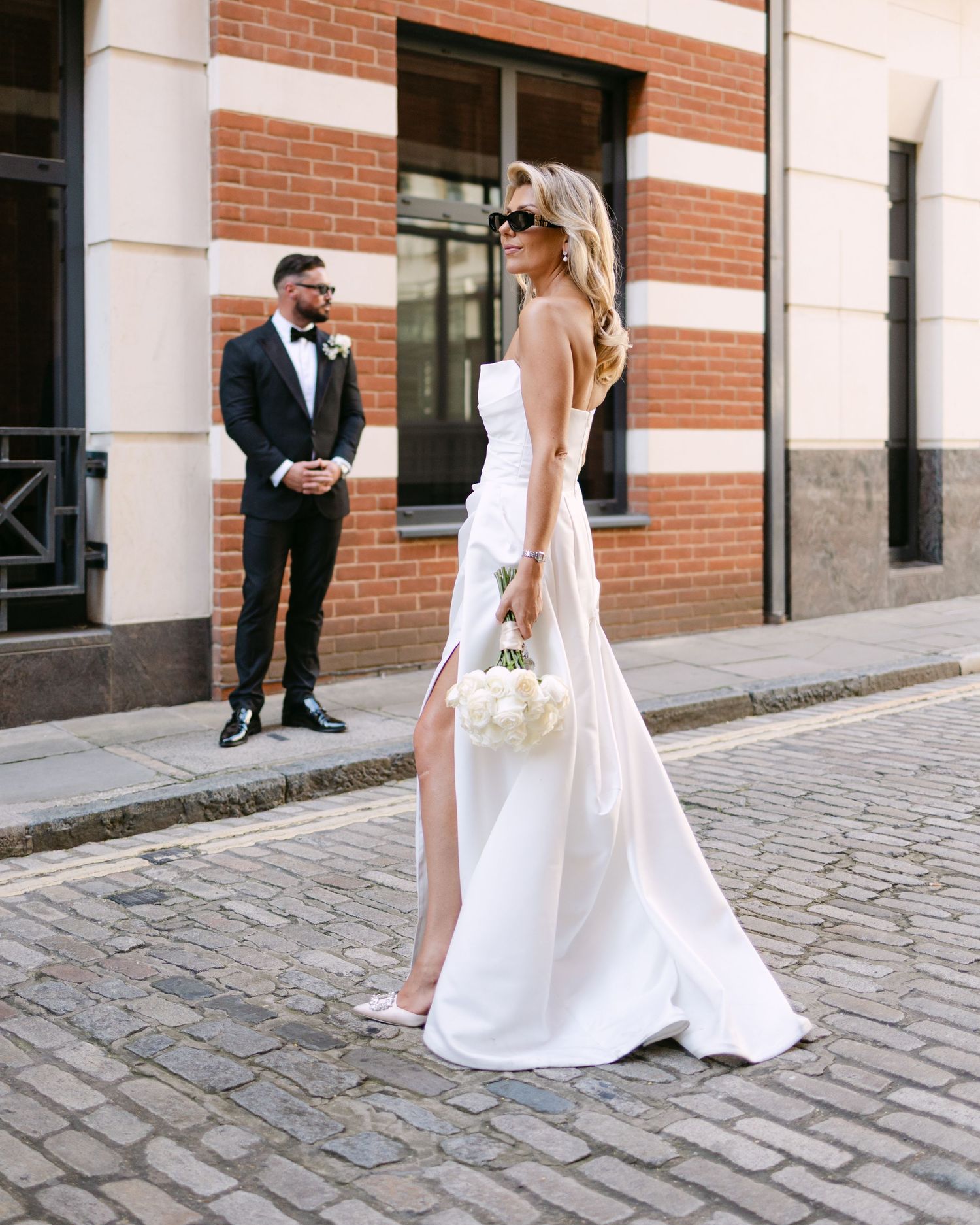 Bride in white strapless gown with slit holds bouquet on cobblestone street as groom in tuxedo watches.