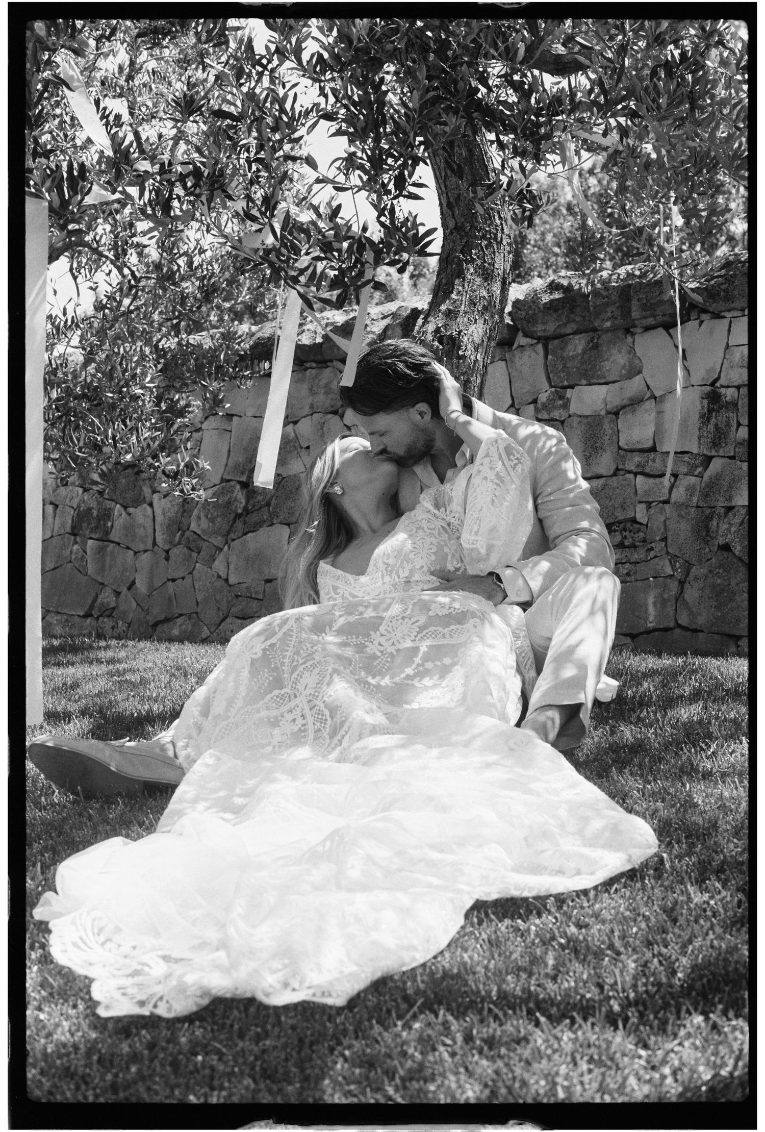 Bride and groom sharing a tender kiss while seated on grass beside a stone wall, romantic black and white photo.