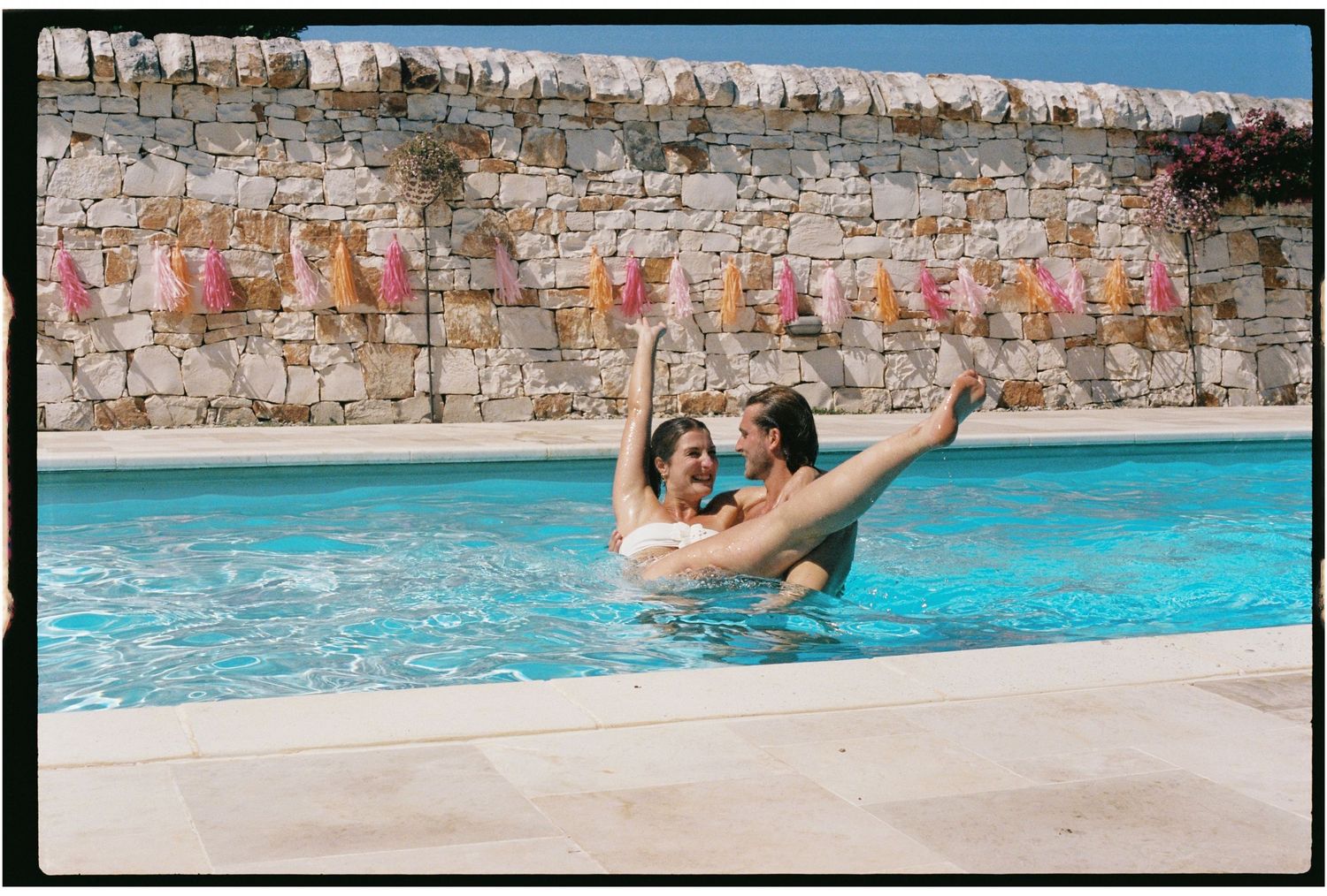 Two women laughing and playing in a sunny outdoor pool with colorful decorations on a stone wall backdrop.