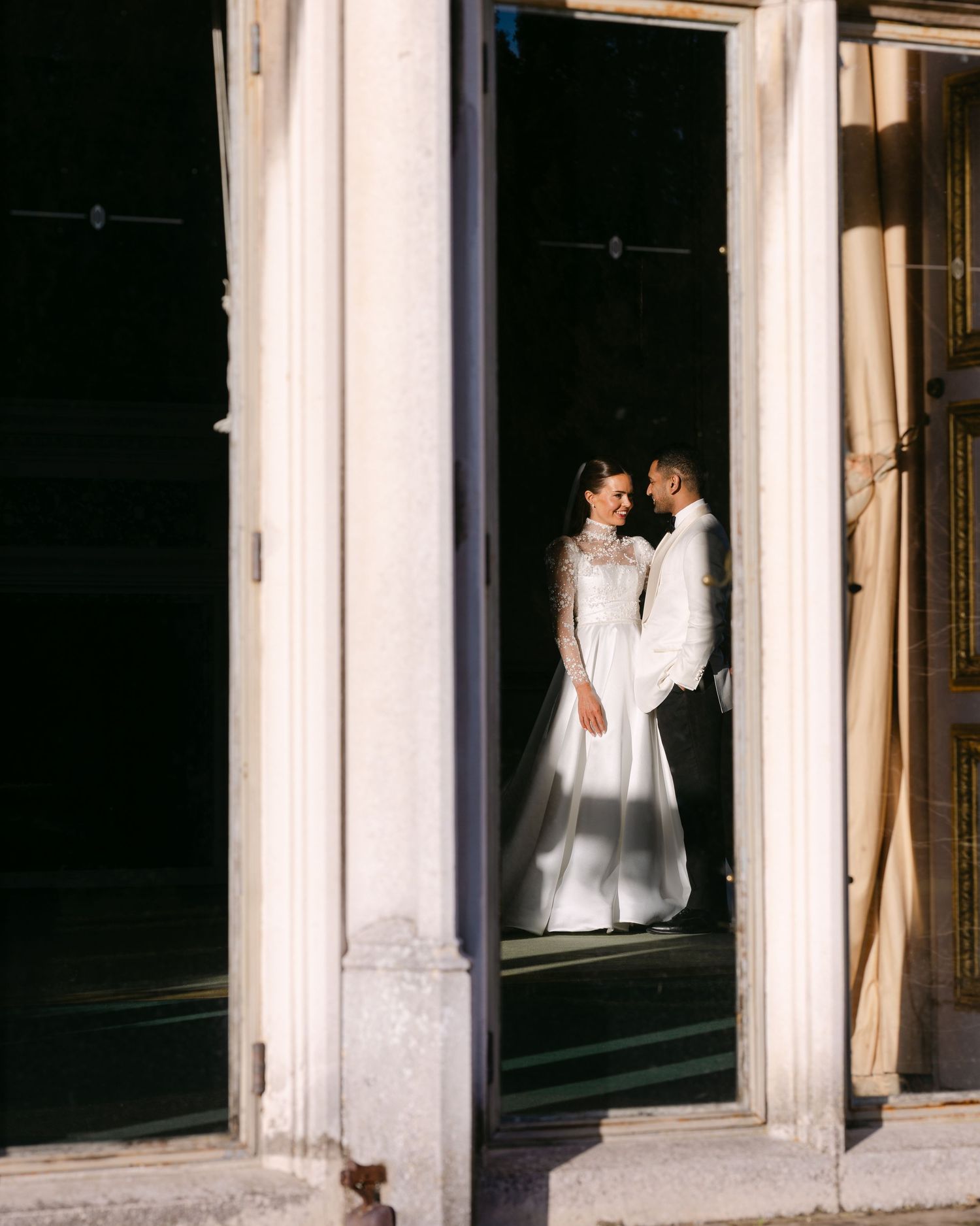Romantic wedding moment captured through window frames in dramatic lighting at Ashridge House, Hertfordshire.