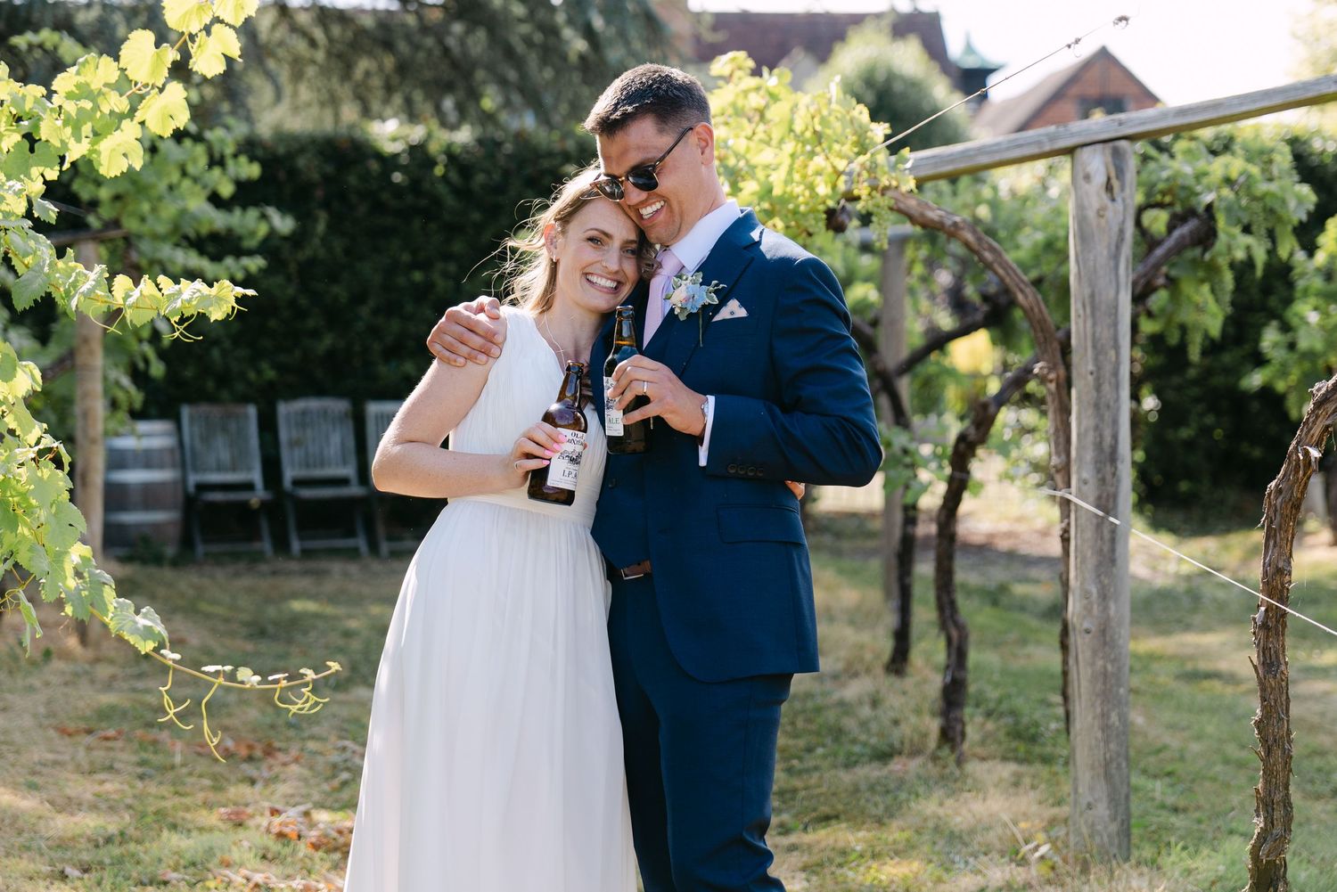 Newlywed couple sharing a champagne bottle outdoors in a vineyard, bride in white dress and groom in navy suit.