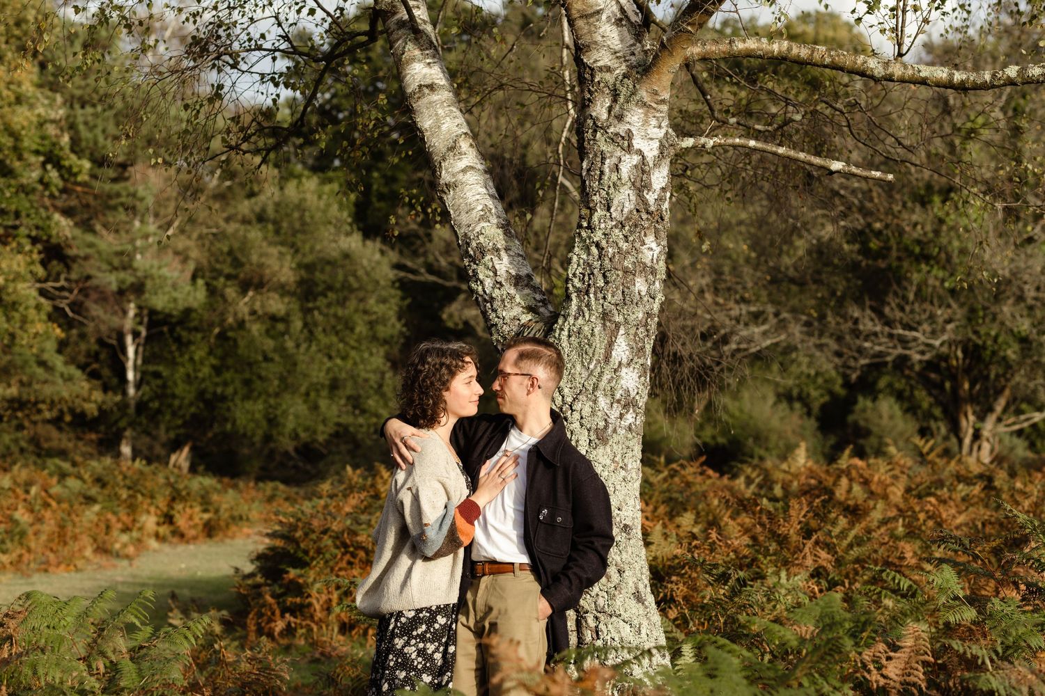 A couple shares intimate moments in a wooded area with autumn trees and filtered sunlight.