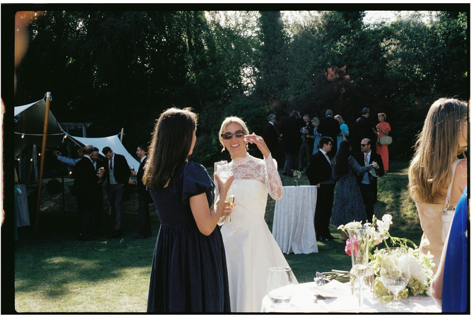Bride in white gown chatting with guests at an outdoor wedding reception on a sunny day.