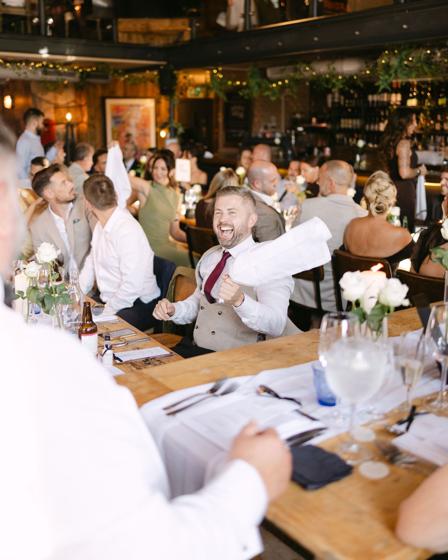 Wedding reception guests celebrate at long wooden tables in rustic restaurant setting.