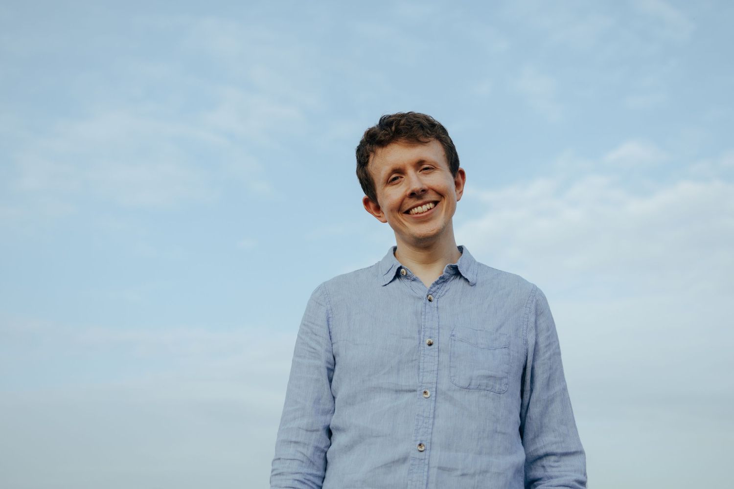 A person in a light blue button-down shirt stands against a cloudy sky.