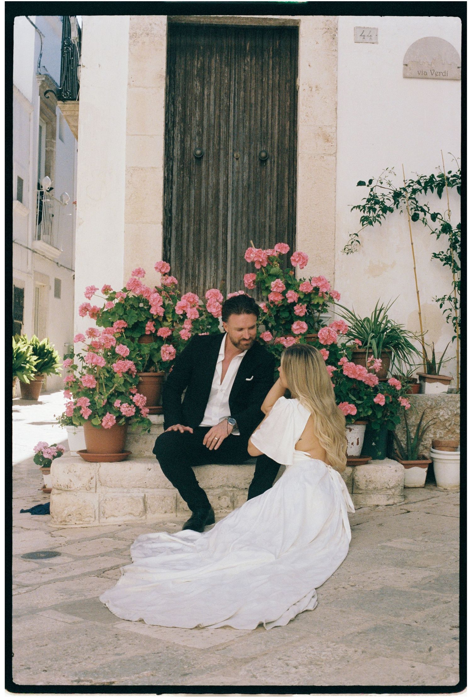 Bride and groom sitting together in front of pink flowers and rustic wooden door in Italy.