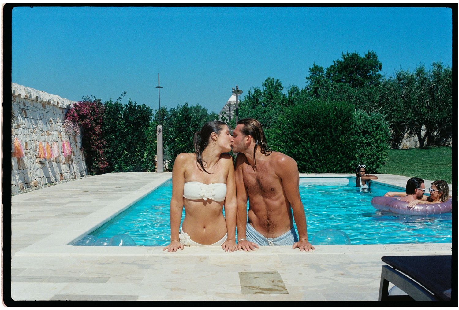A couple sharing a romantic kiss poolside at a sunny outdoor swimming pool surrounded by lush greenery.