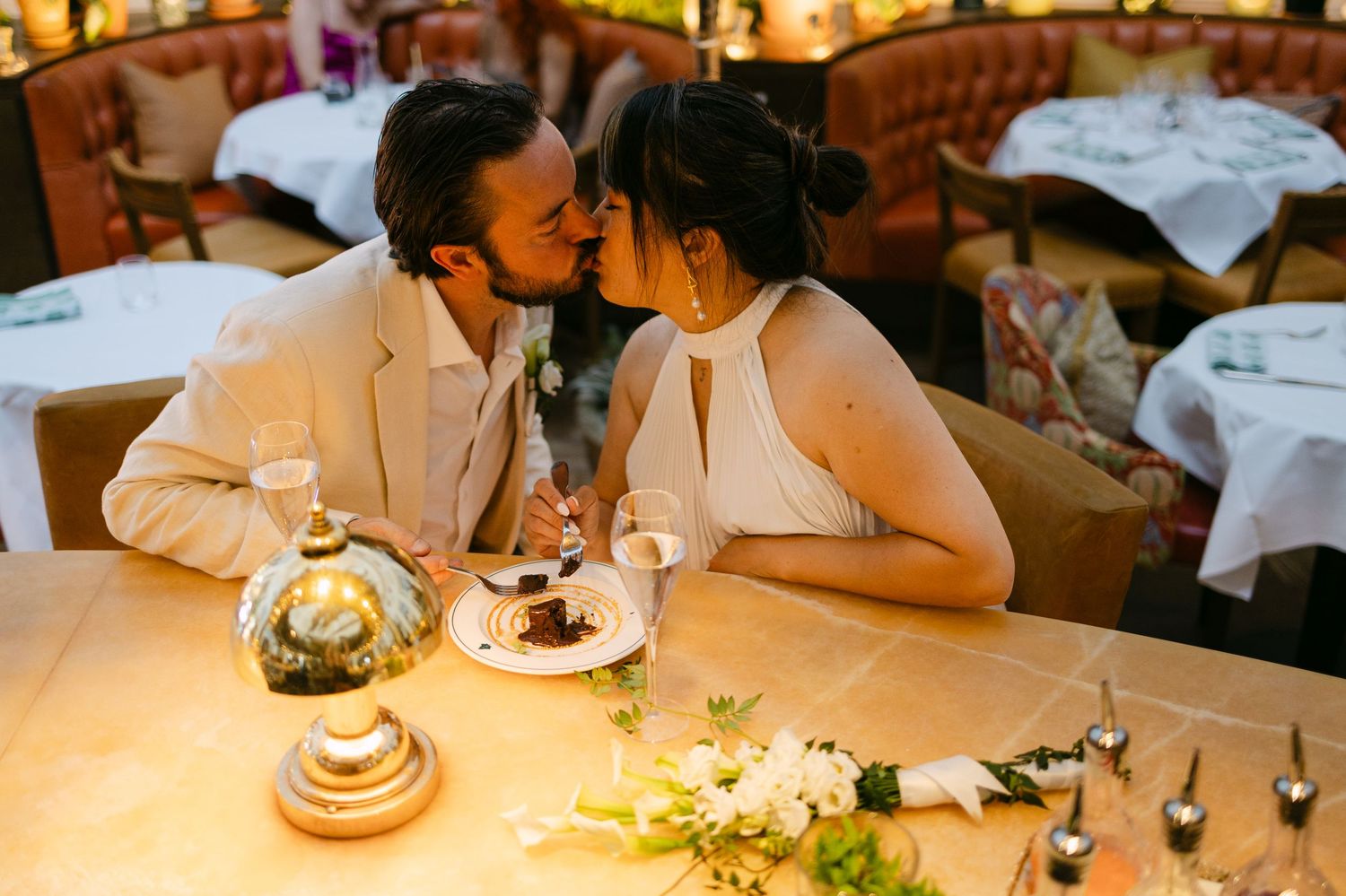 Newlywed couple sharing a romantic kiss at an elegant restaurant table with floral centerpiece.