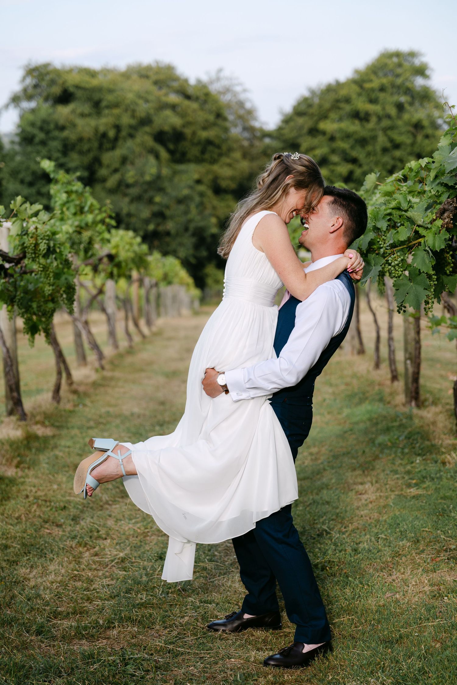 Groom lifting bride in white dress for a romantic kiss in a lush vineyard during their wedding day.