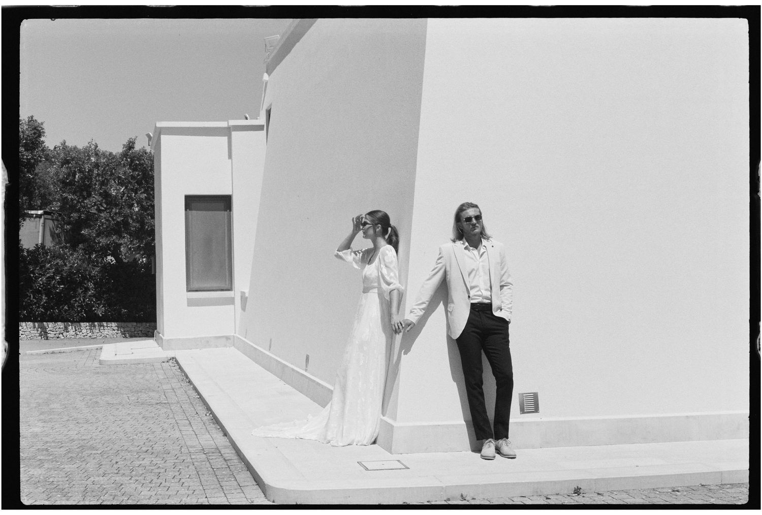 A stylish couple poses against a white building exterior in elegant black and white fashion photography.