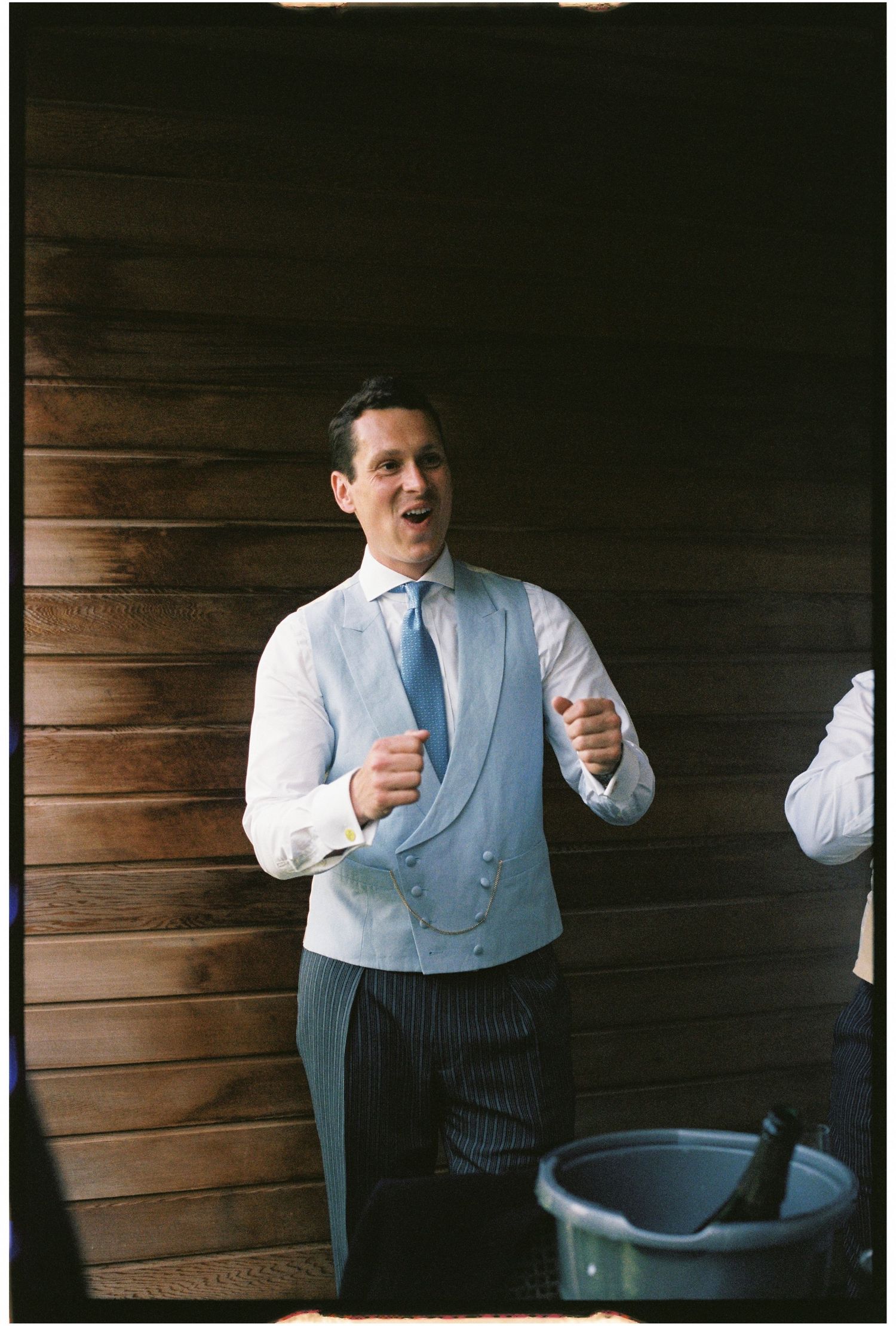Smiling groom in a light blue waistcoat and striped trousers stands against a wooden wall.