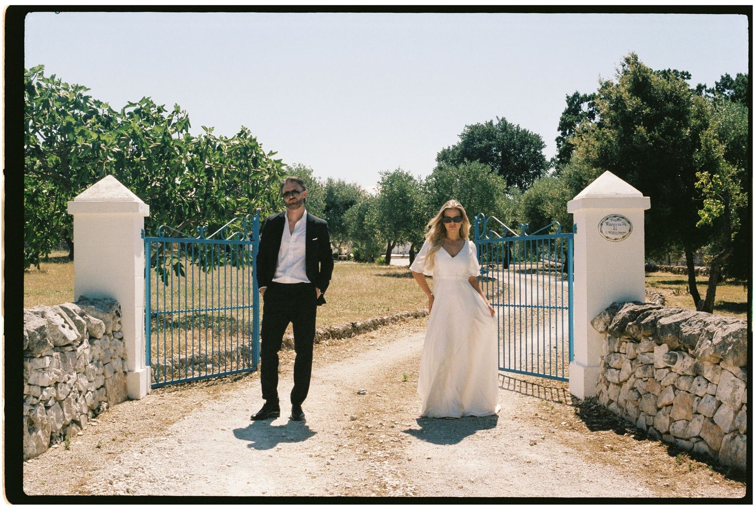 Bride and groom standing at a blue iron gate surrounded by olive trees in Italy.
