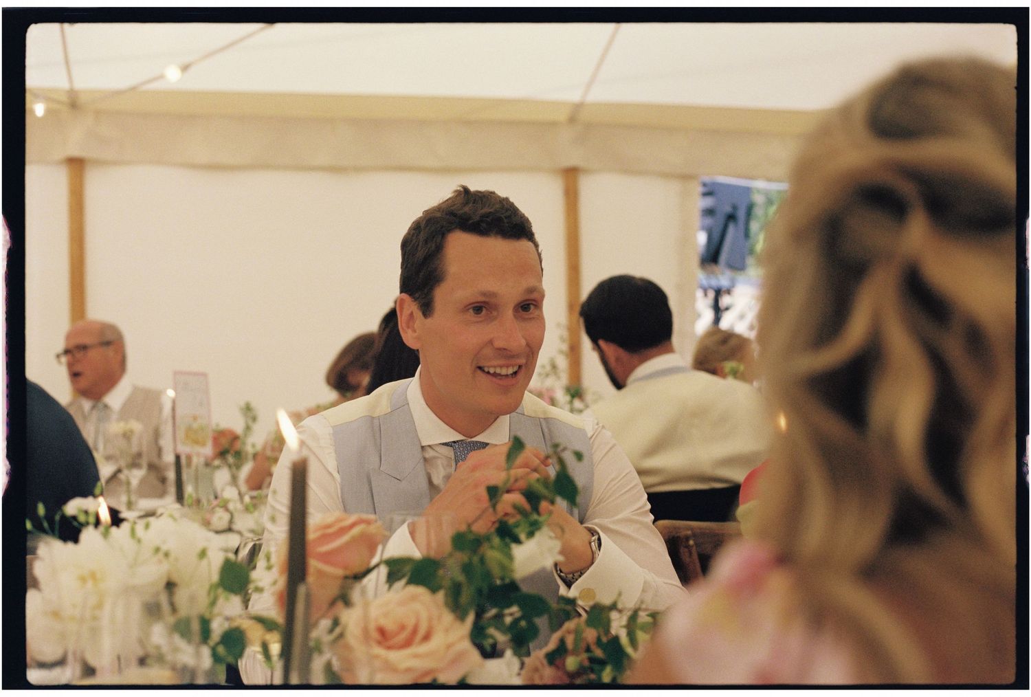 Man in light suit conversing at elegant wedding reception under marquee tent with floral centerpieces.