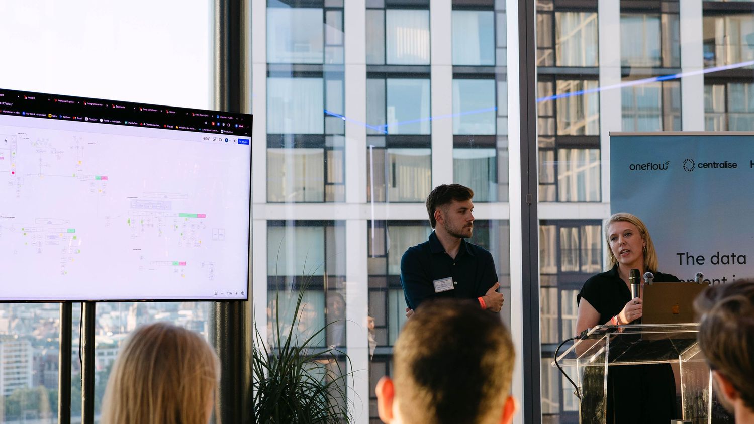 Two presenters speak at a podium during a data conference with a large screen displaying analytics dashboards.