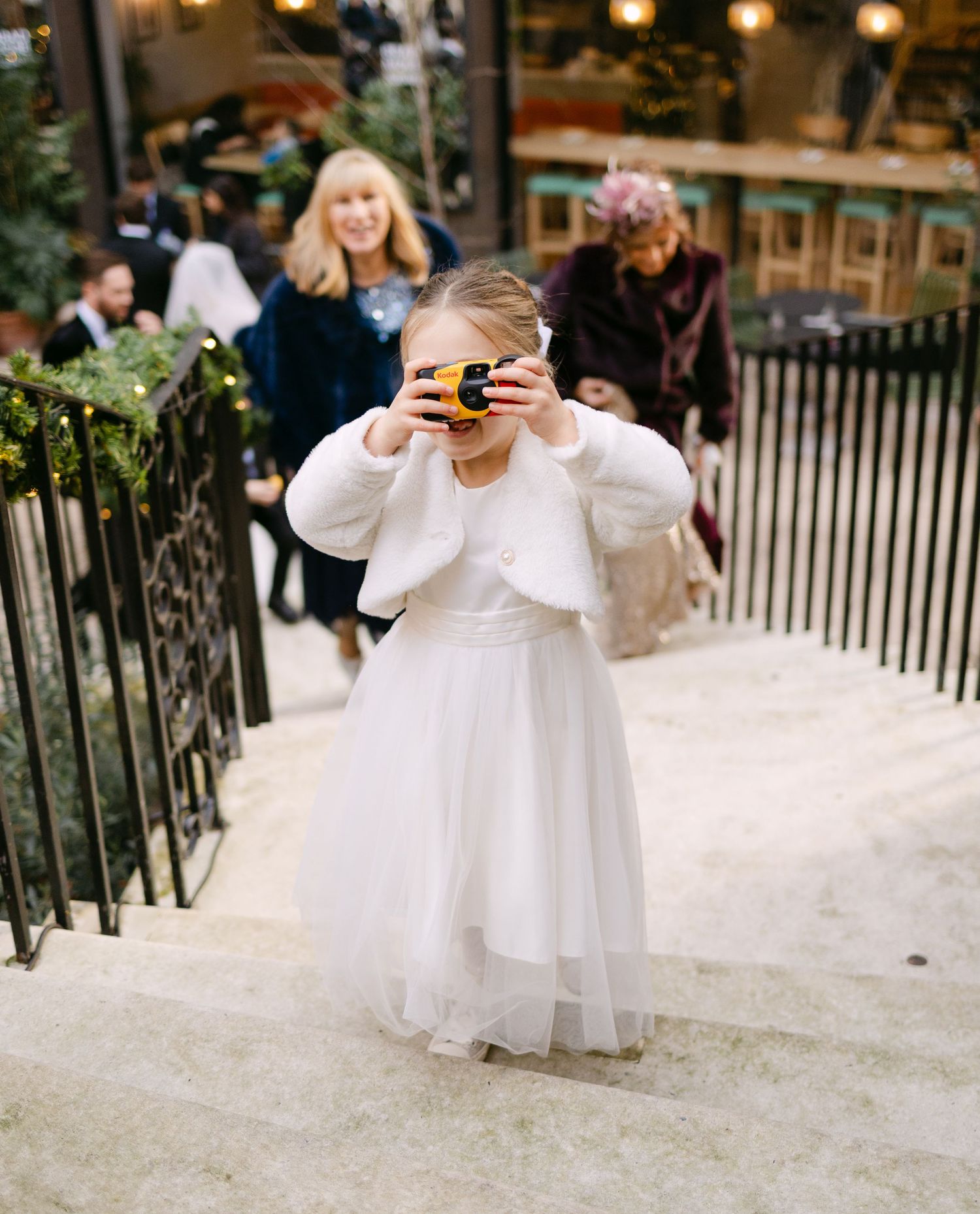 Young flower girls in white dresses play with cameras during wedding festivities.