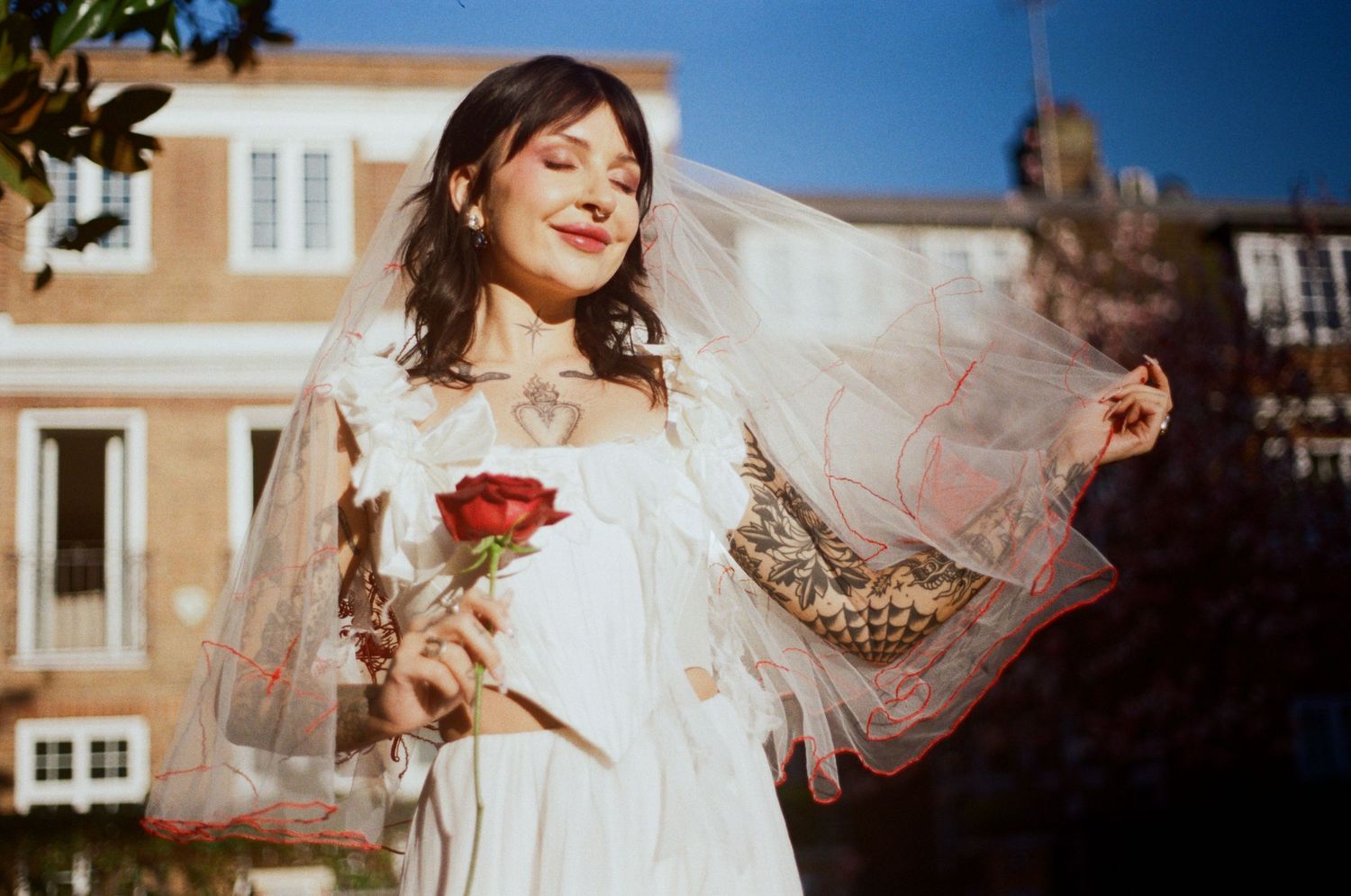 Woman in white dress holding red rose with veil blowing in wind outdoors.