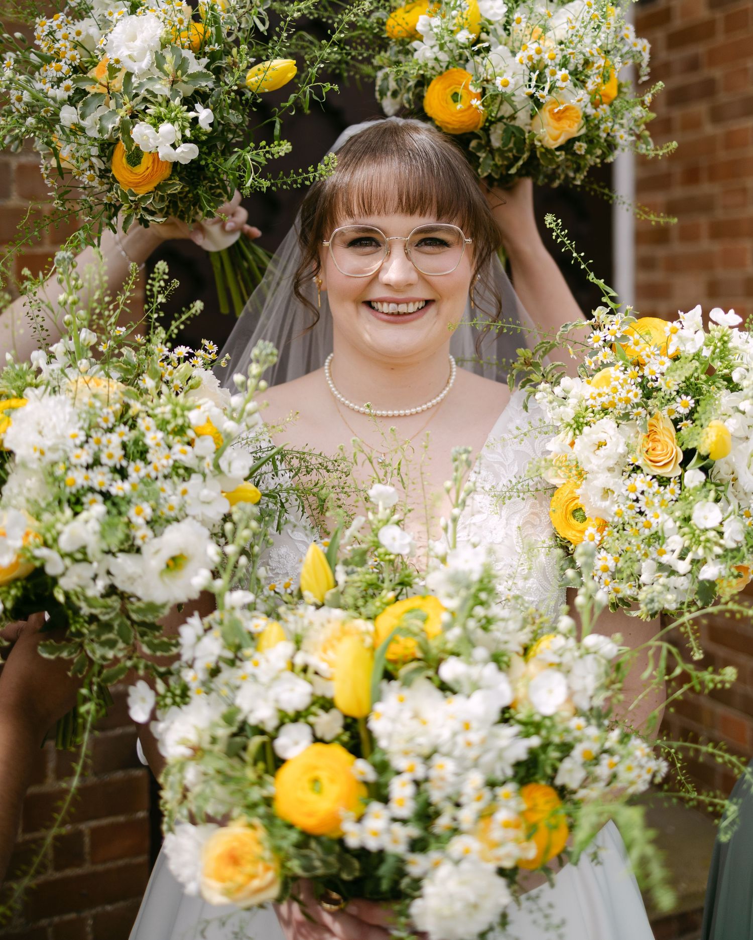 Smiling bride in white gown and veil surrounded by yellow and white floral bouquets on her wedding day.