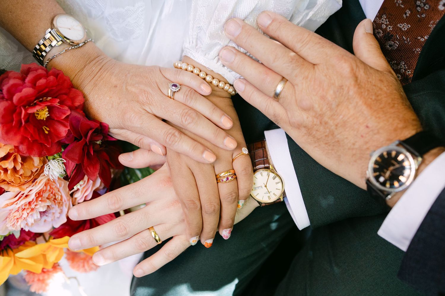 Close-up of wedding hands displaying rings, watches, and jewelry beside vibrant red and orange floral bouquet.