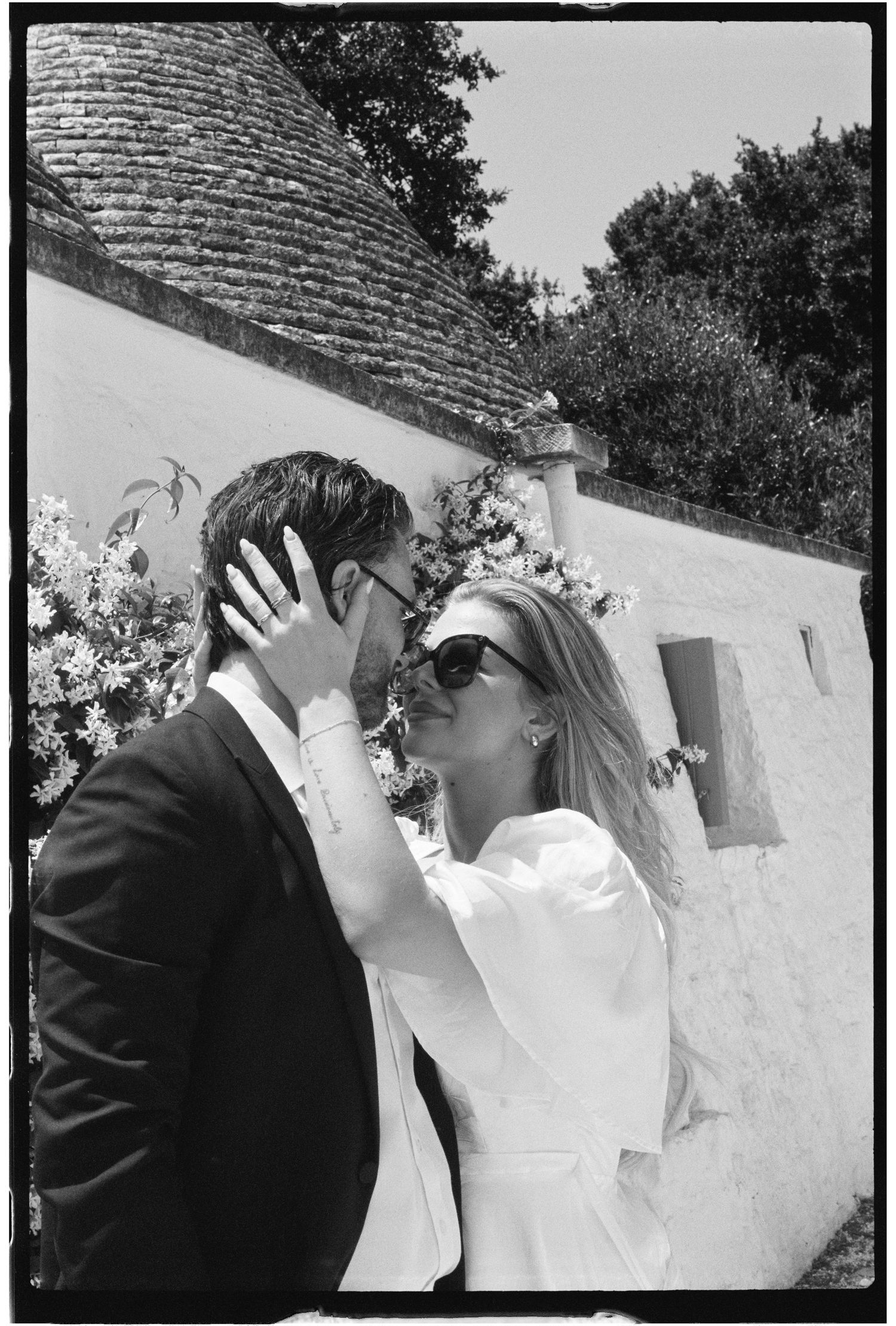 Black and white photo of a bride and groom sharing a romantic kiss outdoors near a rustic white wall.