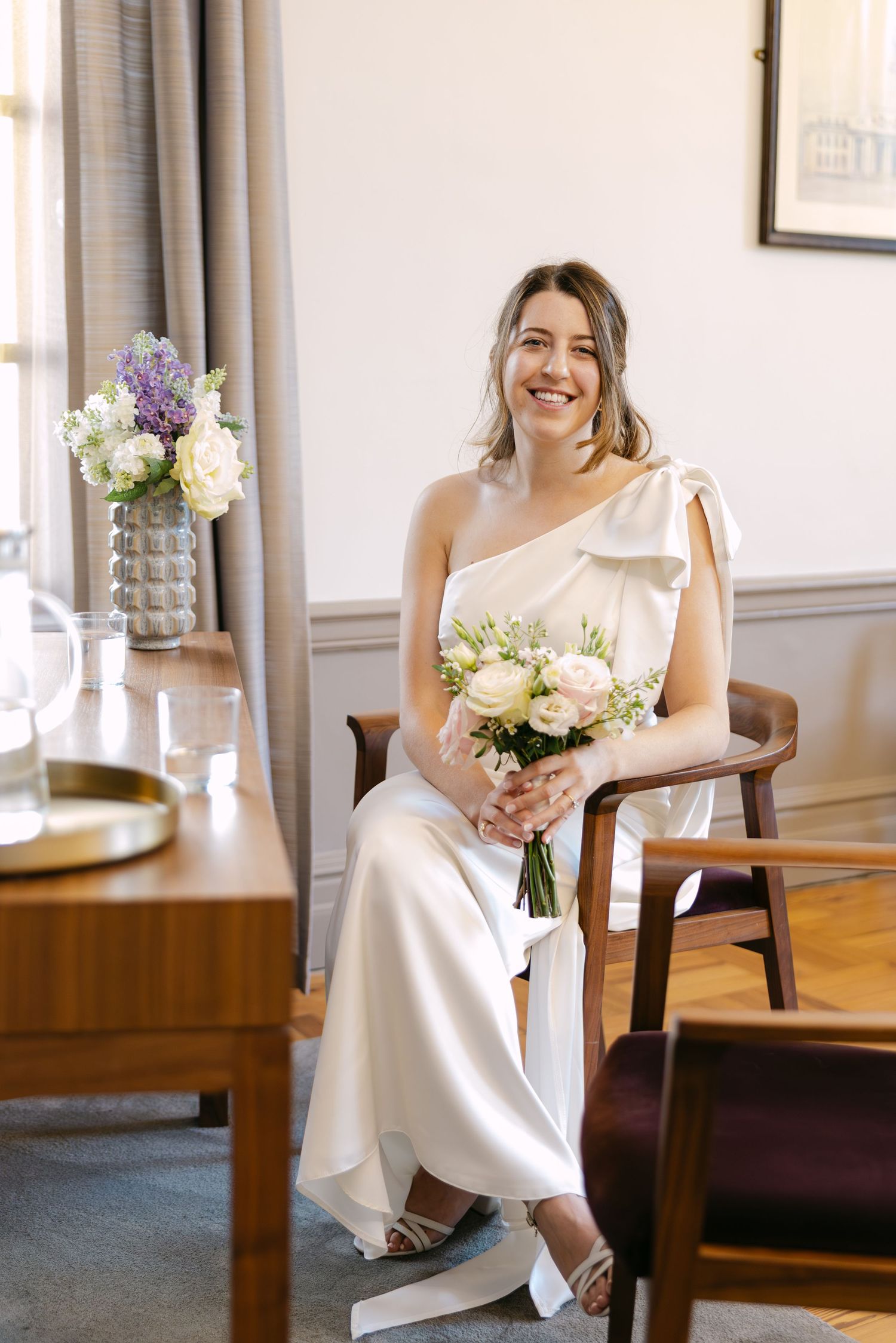 A bride in a white dress sits on a wooden chair holding a bouquet of flowers in a sunlit room at The Old Marylebone Town Hall.