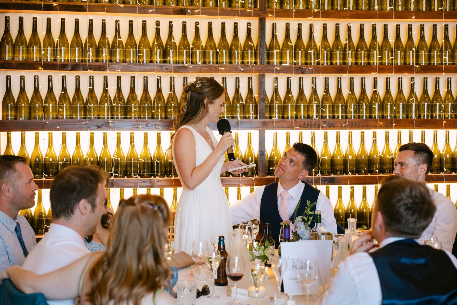 Wedding guest delivers a speech at a winery reception, with glowing wine bottle wall display behind the head table.