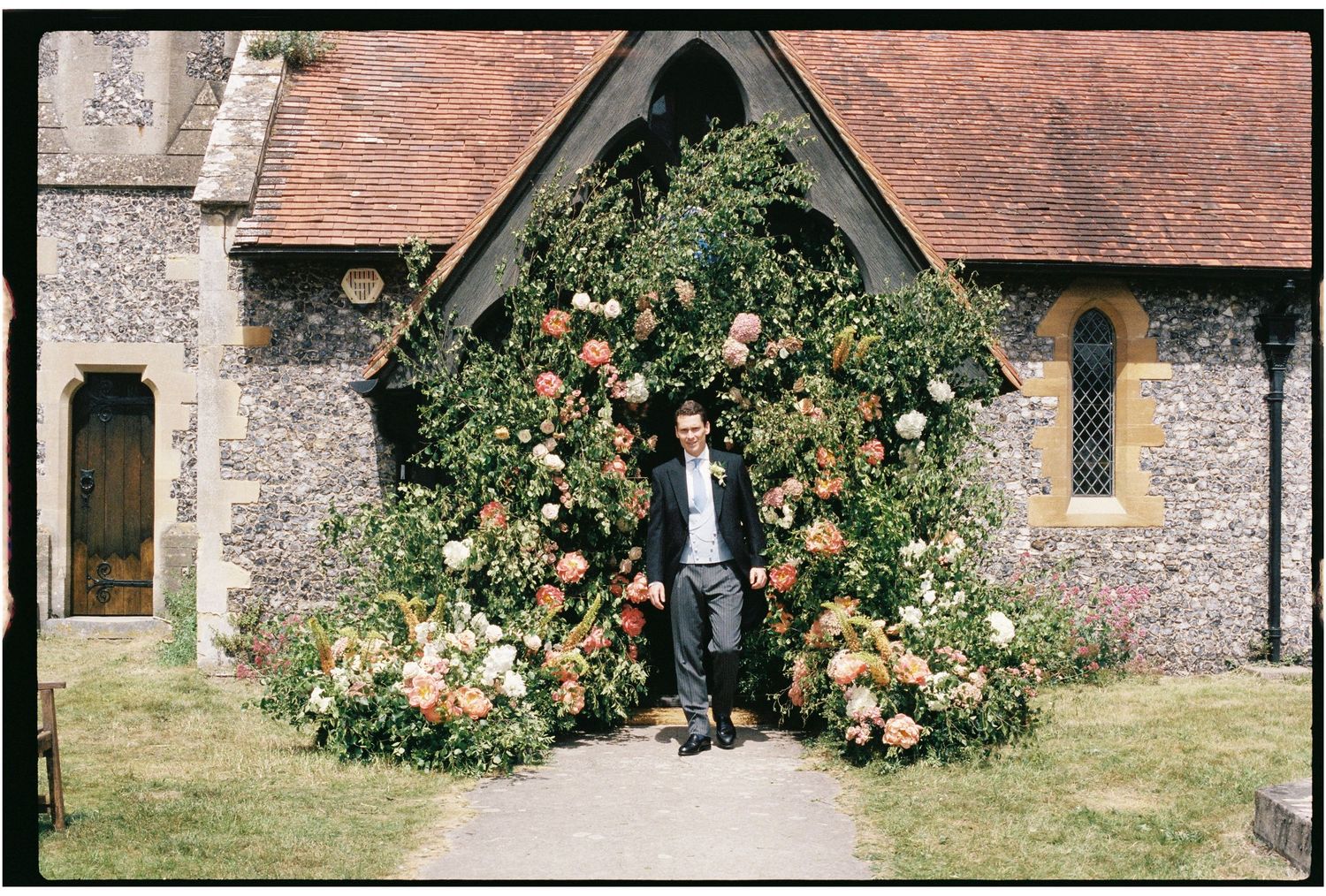 Groom standing beneath a stunning floral arch of roses at the entrance of a traditional English stone church.