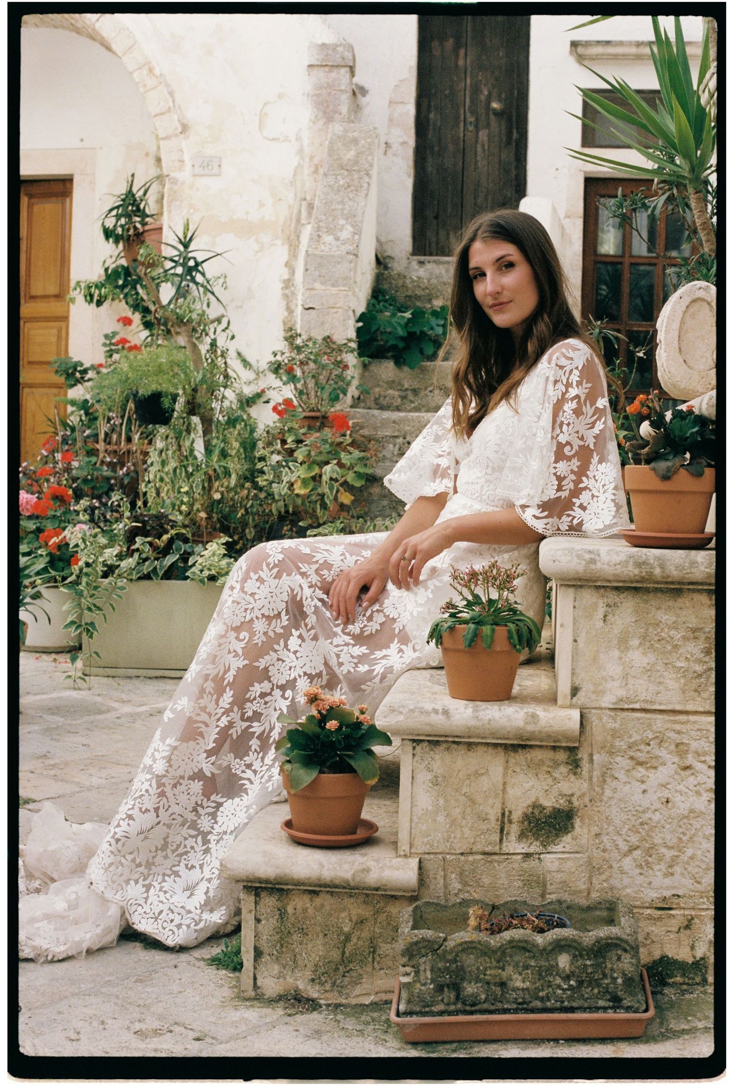 Bride in sheer lace gown seated on stone steps surrounded by terracotta pots and lush greenery in a rustic courtyard.