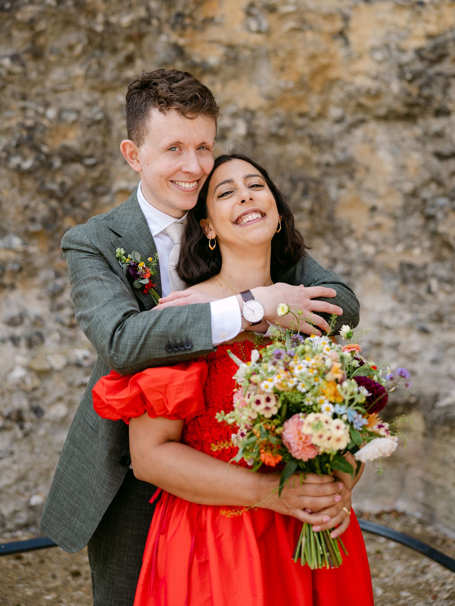 Happy couple shares joyful moments during their wedding photoshoot by a stone wall.
