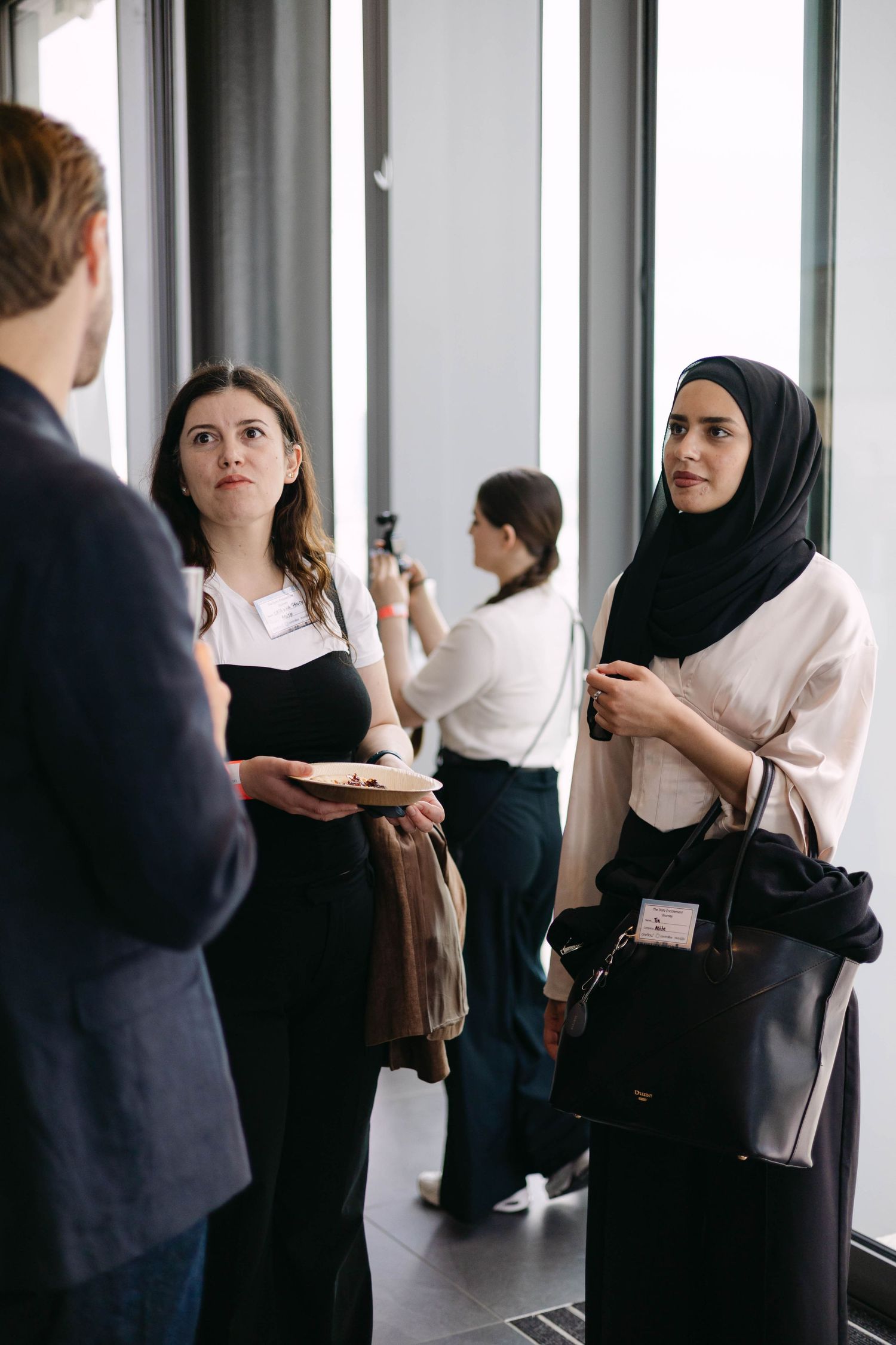 Diverse women networking at a professional conference, one in hijab, another holding a plate, conversing indoors.