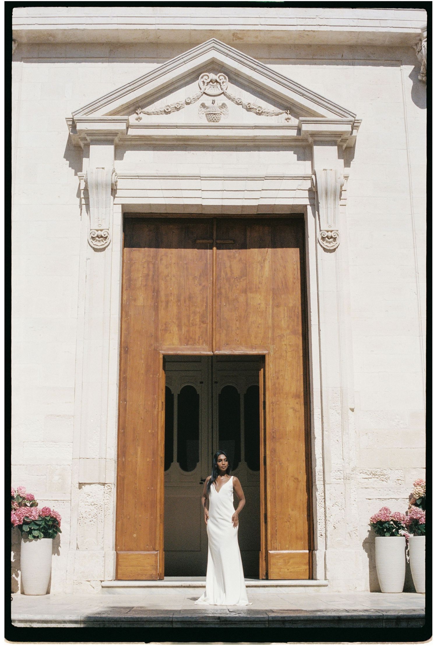 Bride in elegant white gown standing in grand church doorway with pink floral arrangements.